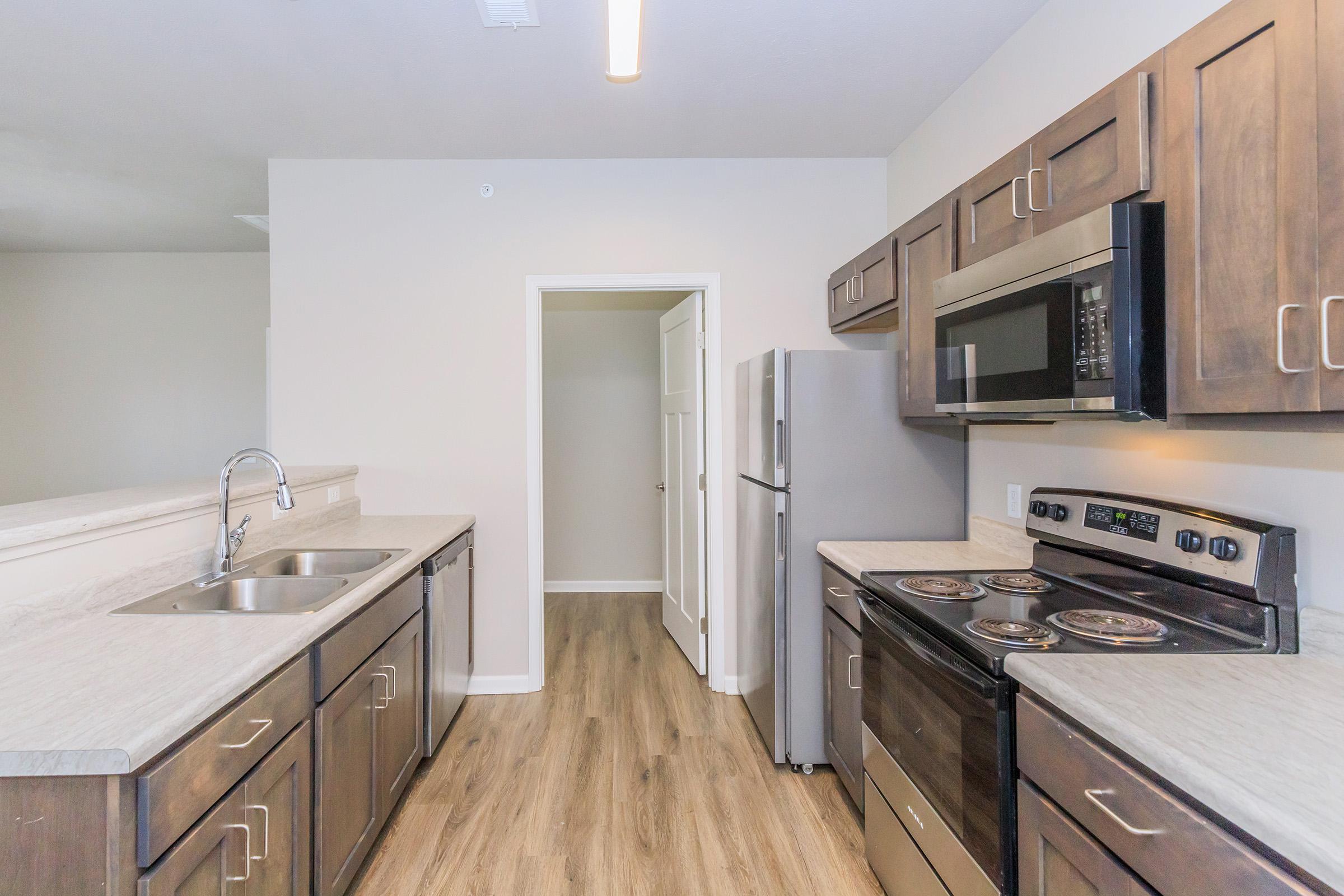 Modern kitchen with dark wooden cabinets, stainless steel appliances, and a spacious countertop. Features a double sink, electric stove, and a microwave. Light-colored walls and laminate flooring create a bright, inviting atmosphere. A doorway leads to another room.