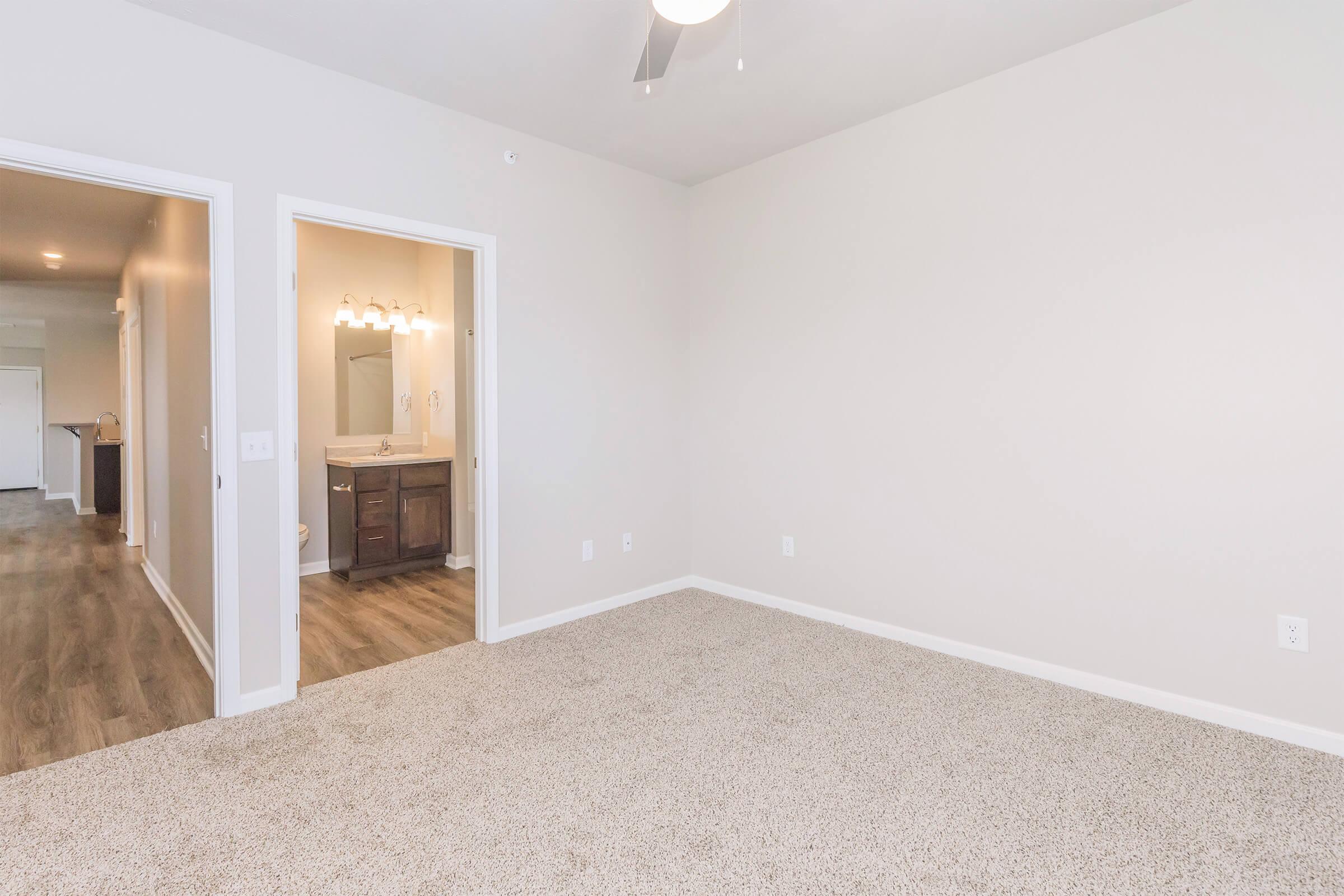 A spacious, unfurnished room with light-colored walls and carpeting. It features a ceiling fan and an open doorway leading to a bathroom with vanity lights. The adjacent doorway suggests a layout connecting to another area, creating a bright and inviting atmosphere.