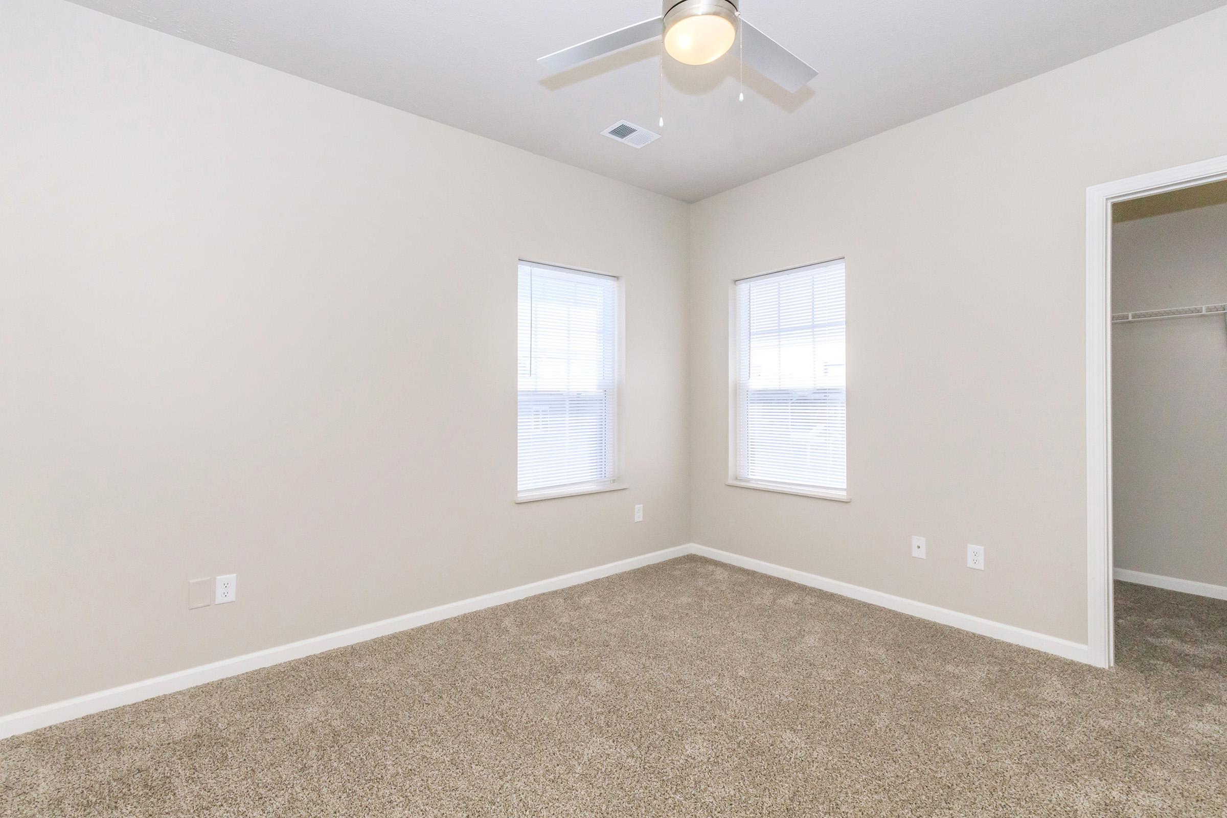 A spacious, empty room featuring light beige walls and a carpeted floor. It has two windows allowing natural light and a ceiling fan. A closet is visible on the right side, contributing to the room's simple and open aesthetic.