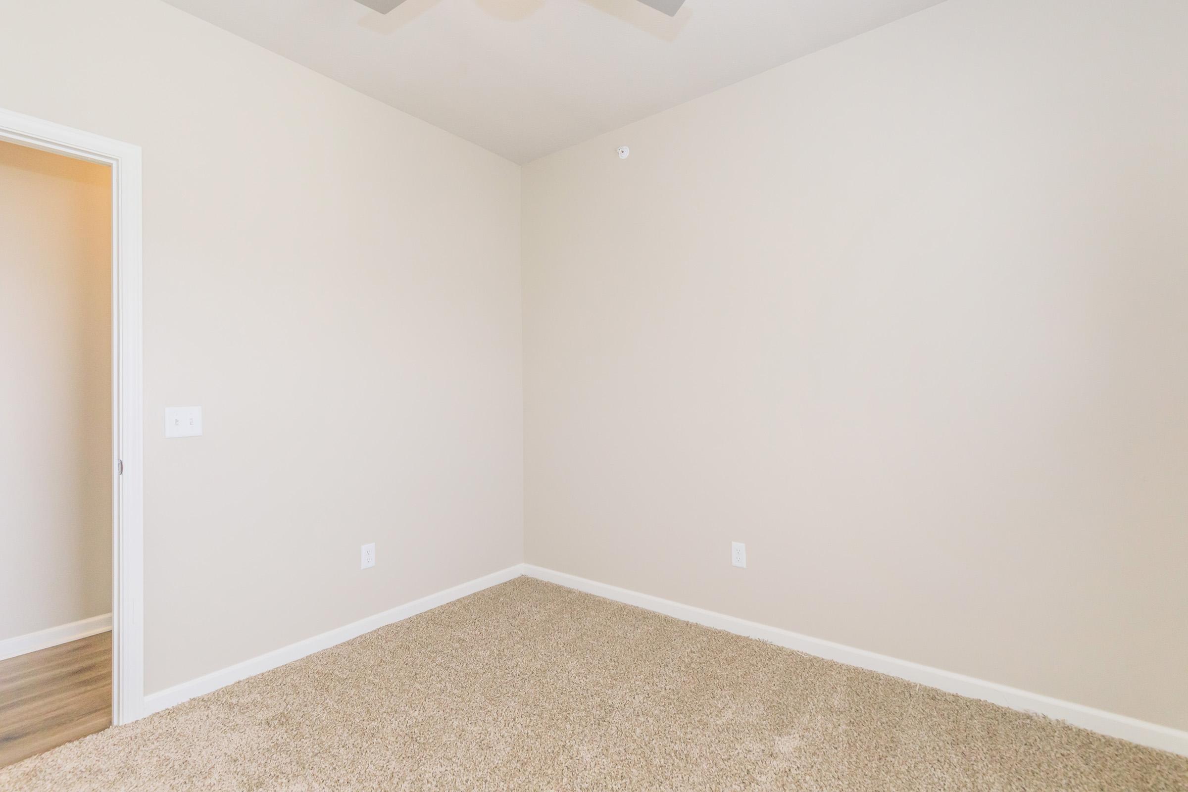 A spacious, empty room with beige walls and a light-colored carpet. There is a ceiling fan and a doorway on the left side leading to another area. The room is well-lit and has no furnishings, creating a clean and minimalistic aesthetic.