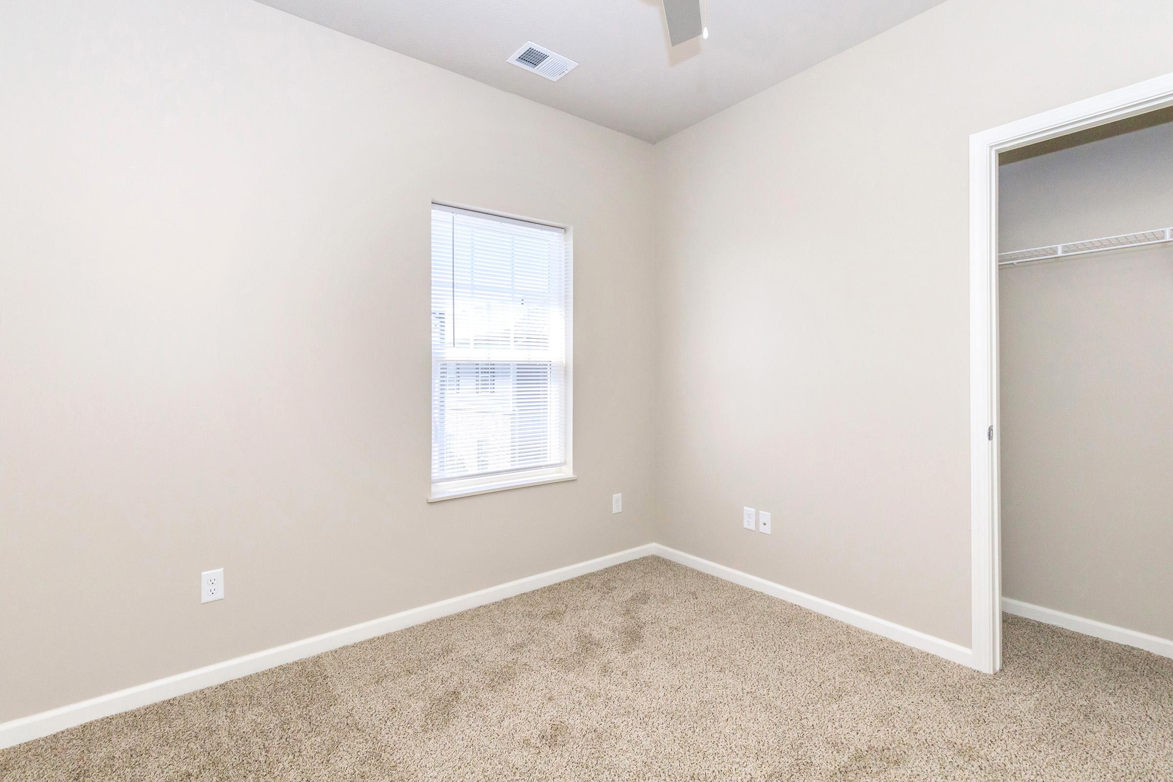 A vacant room with light beige walls and carpeted flooring. There is a window with blinds, allowing natural light to enter. The room features a ceiling fan and a closet door on the right. The overall space is clean and minimalistic, ideal for personal customization.