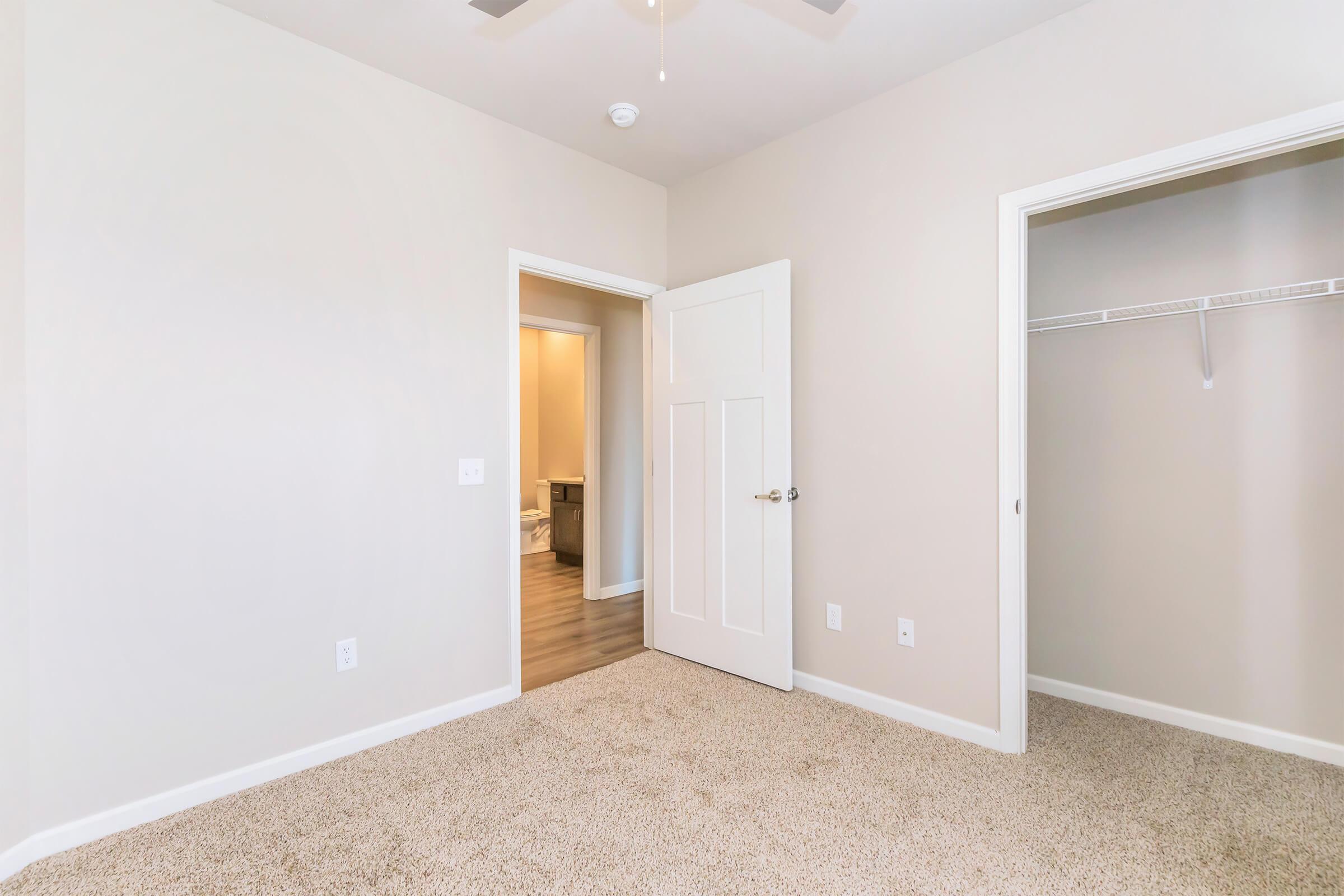 A well-lit room featuring beige walls and carpet flooring. There is a closed door leading to a bathroom, and another door that opens into a closet with a shelf. A ceiling fan is visible above, adding to the airy ambiance of the space. The overall look is clean and modern.