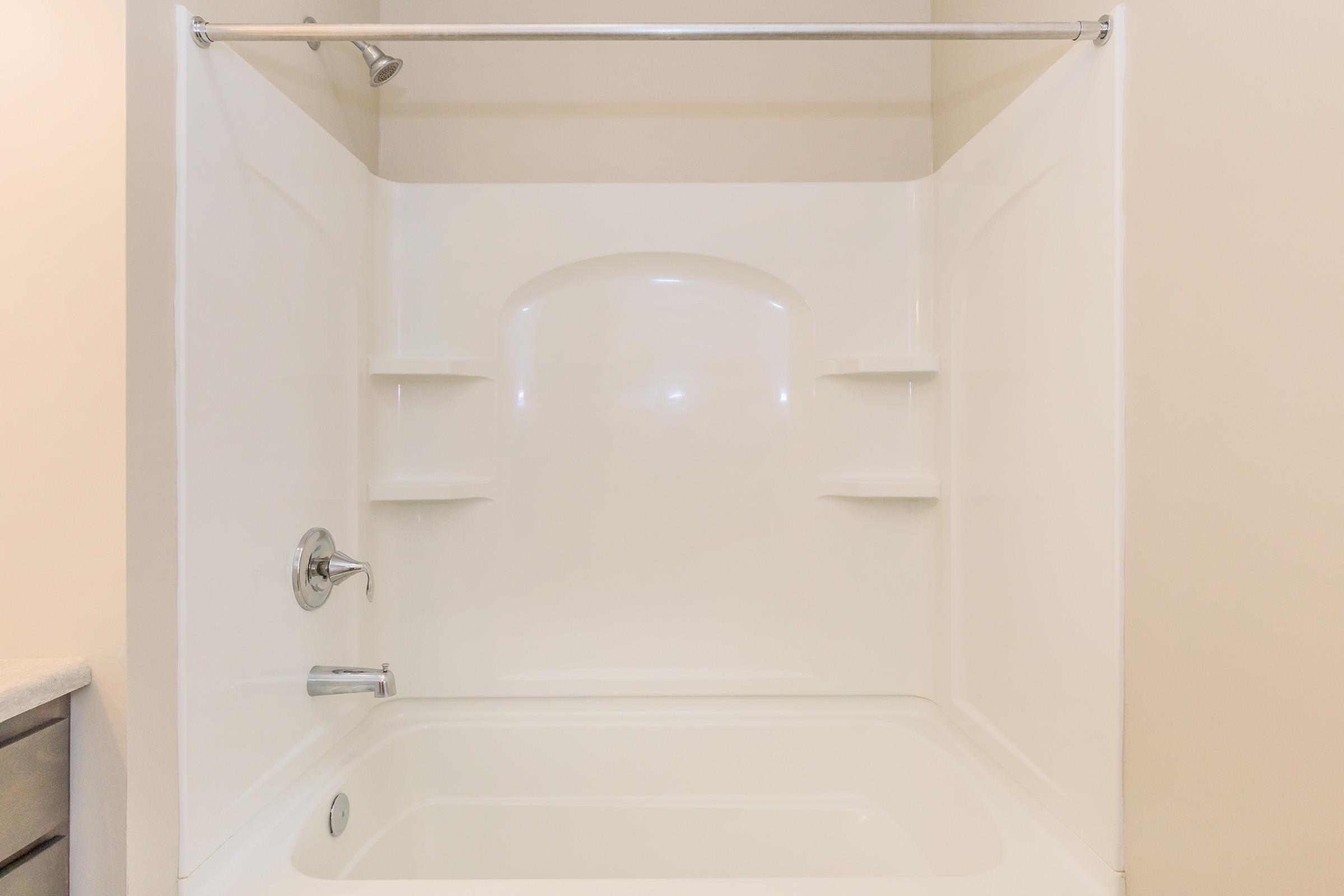 A clean, white bathtub with an attached shower. The surrounding walls are also white, and there are built-in shelves on either side for toiletries. The design is modern and minimalistic, with a chrome faucet and showerhead. The overall appearance is bright and well-maintained.