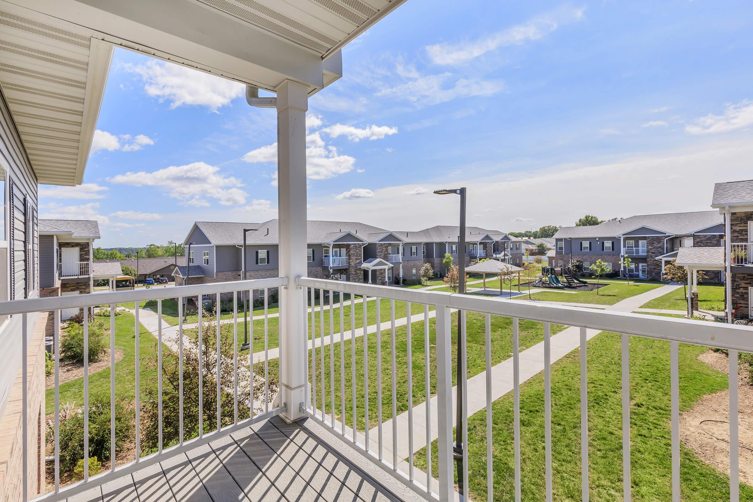 View from a balcony overlooking a well-maintained residential community. Neat sidewalks meander through green lawns, with several two-story buildings in the background under a clear blue sky with a few clouds.