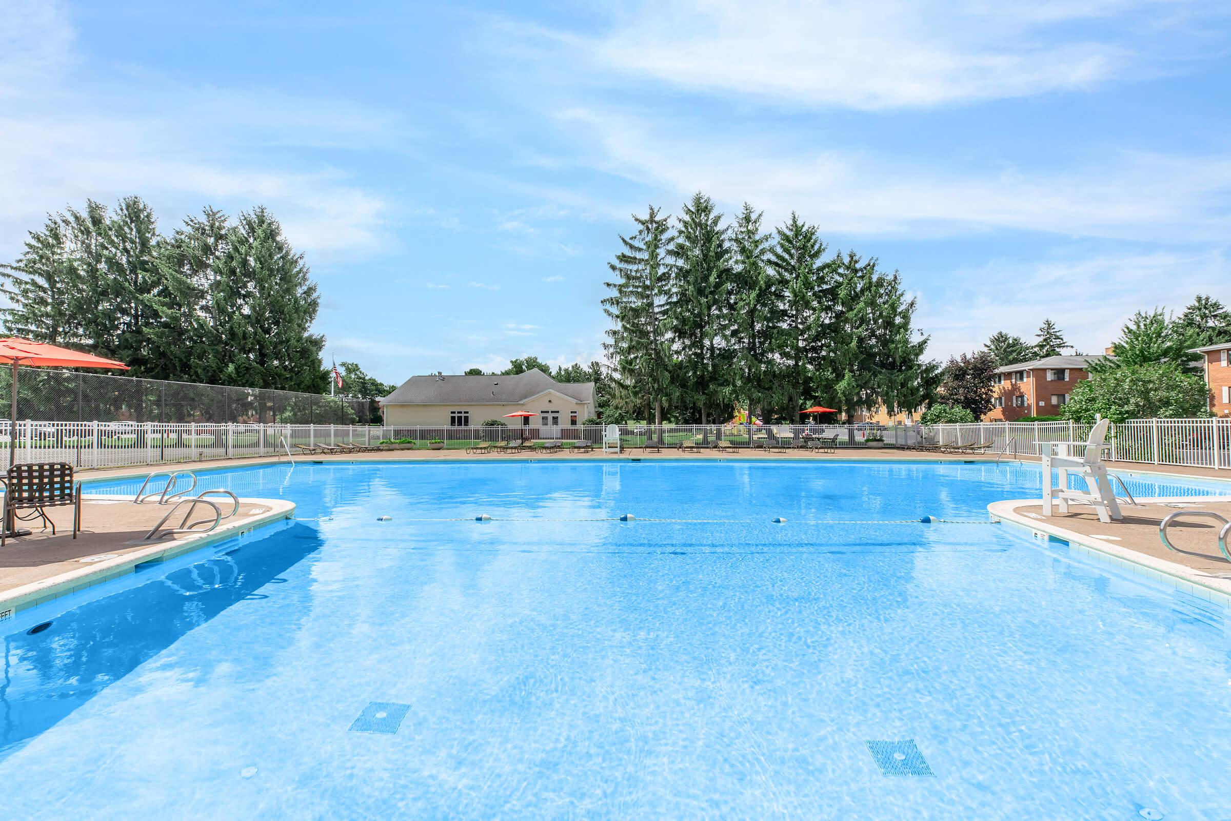 A large outdoor swimming pool surrounded by lounge chairs, with a clear blue sky and green trees in the background. The pool area features a spacious deck and a nearby building visible in the distance.