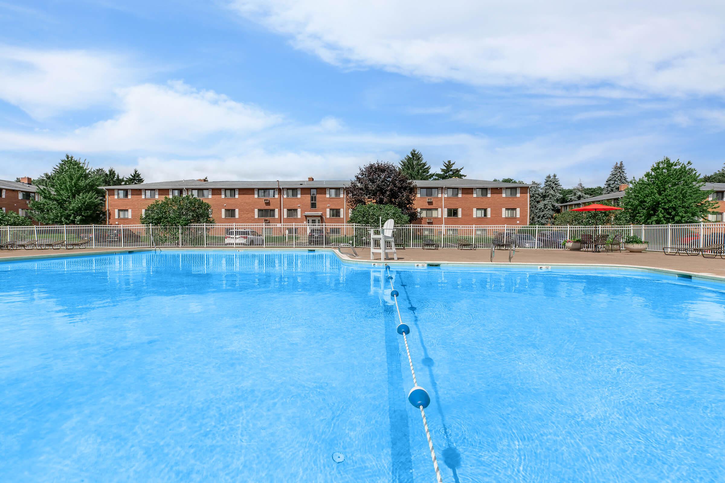 A clear blue swimming pool surrounded by a fence, with a few nearby trees and an apartment building in the background under a partly cloudy sky.