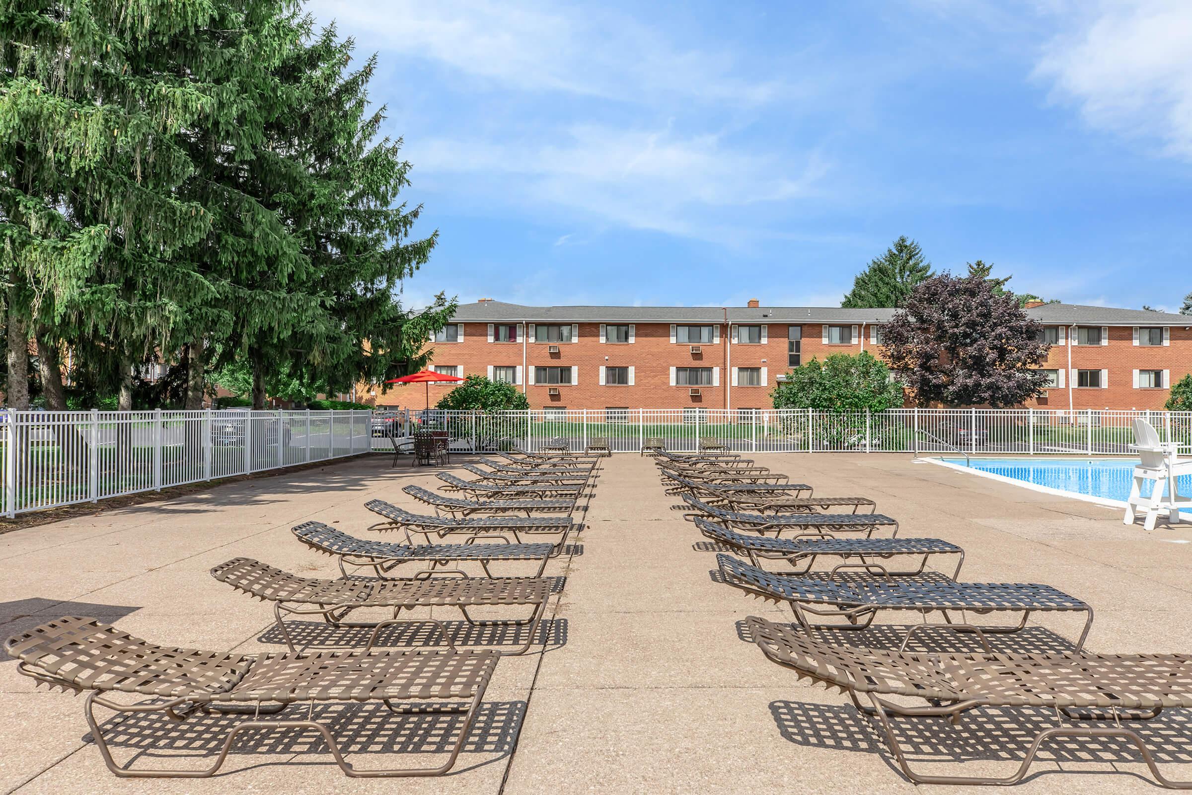 A clear blue sky above a swimming pool area lined with multiple brown lounge chairs arranged in neat rows, surrounded by trees and a brick building in the background.