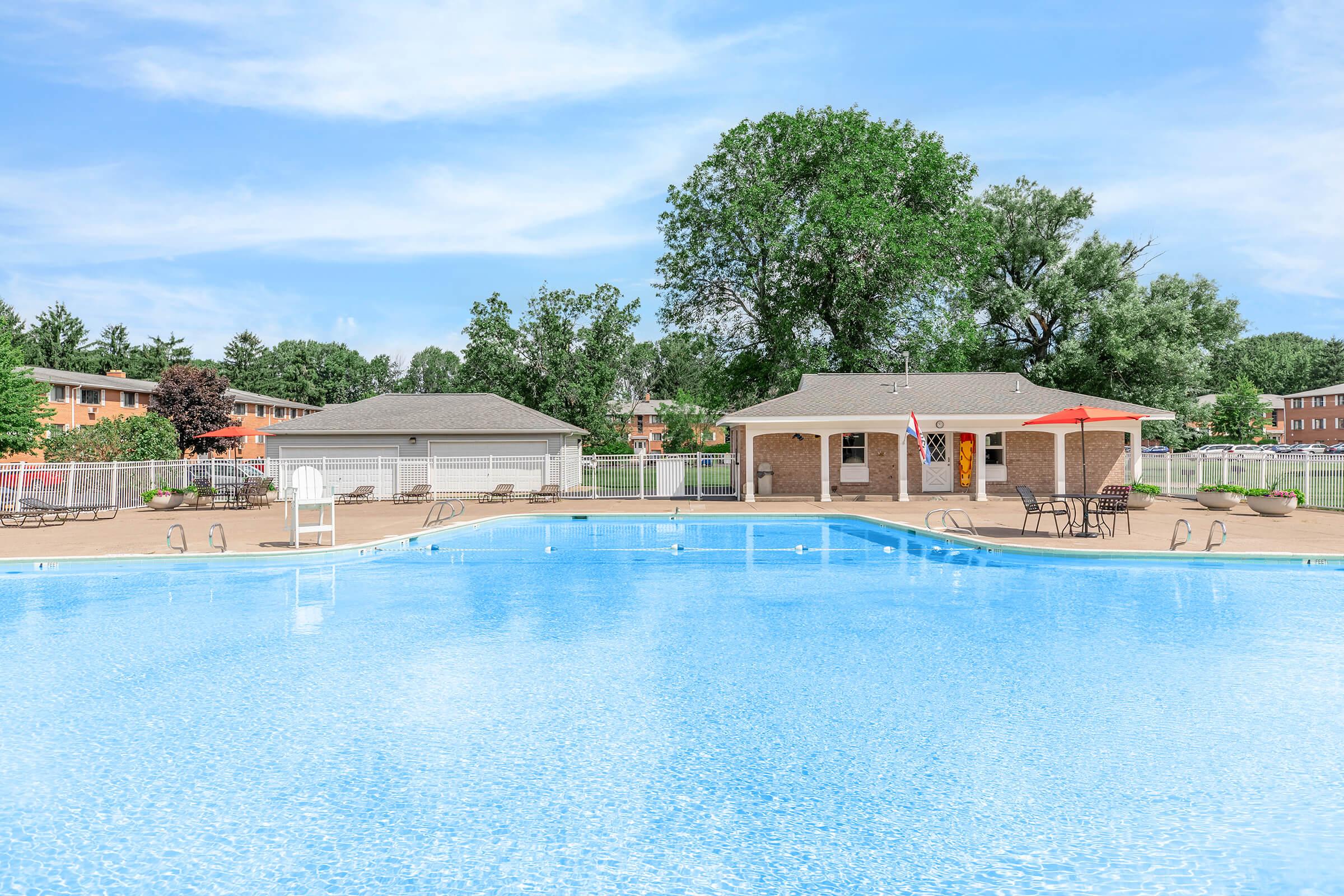 A clean, blue swimming pool surrounded by a light brown patio, with lounge chairs and tables set up. In the background, there is a small brick building with an American flag and a red umbrella. Green trees and residential buildings are visible in the distance under a clear blue sky.