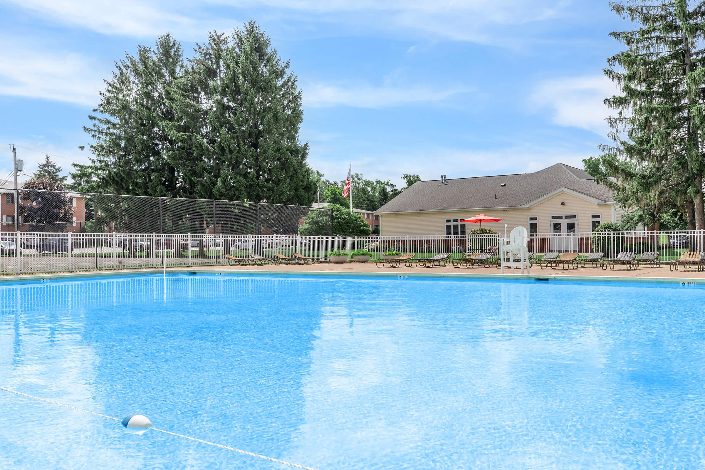 A clear blue swimming pool surrounded by lounge chairs, with a fence in the background. Tall trees and a building are visible on the right side, under a bright blue sky with some clouds.