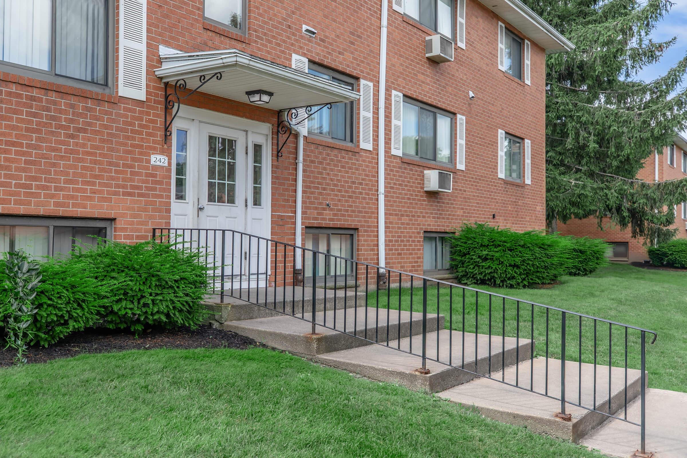 Exterior view of an apartment building featuring a brick façade, a set of stairs leading to a white double door entrance, and landscaped greenery on either side.