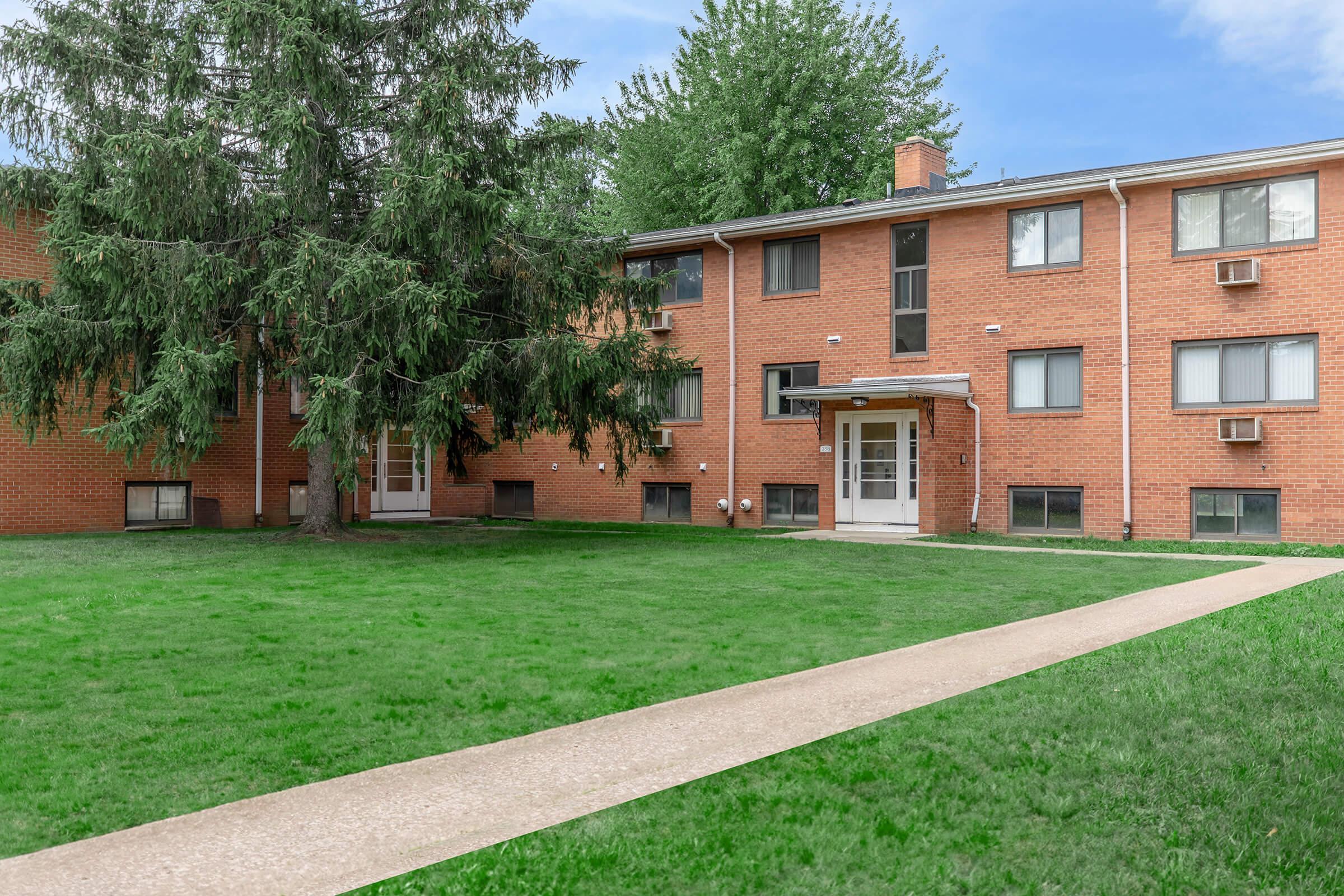 A brick apartment building with multiple windows and a grassy lawn area. A paved walkway leads to the entrance, which is framed by a small overhang. Large trees are visible in the surrounding area.