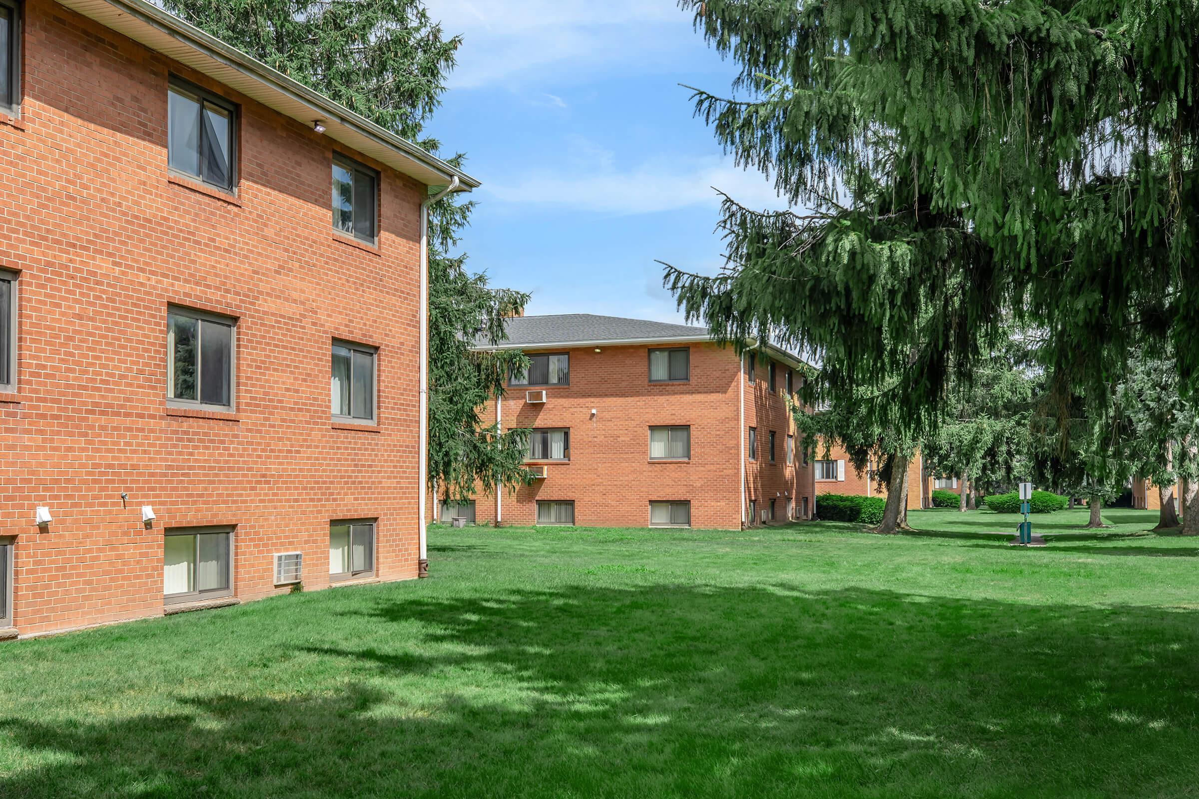 A scene featuring multiple brick apartment buildings surrounded by green grass and trees under a clear blue sky.