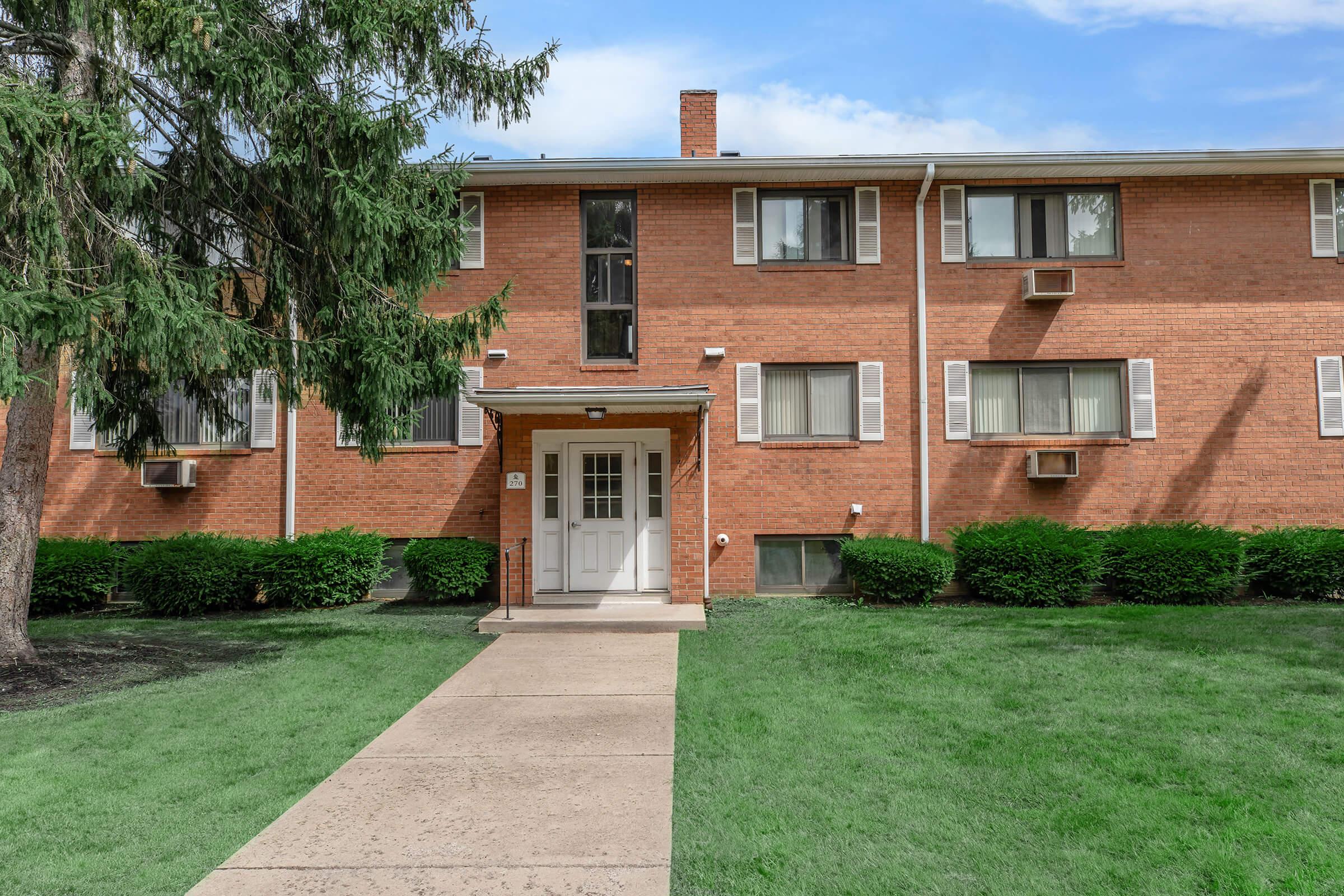 A brick apartment building with multiple windows and air conditioning units, surrounded by green grass and shrubs, and a concrete walkway leading to the entrance.