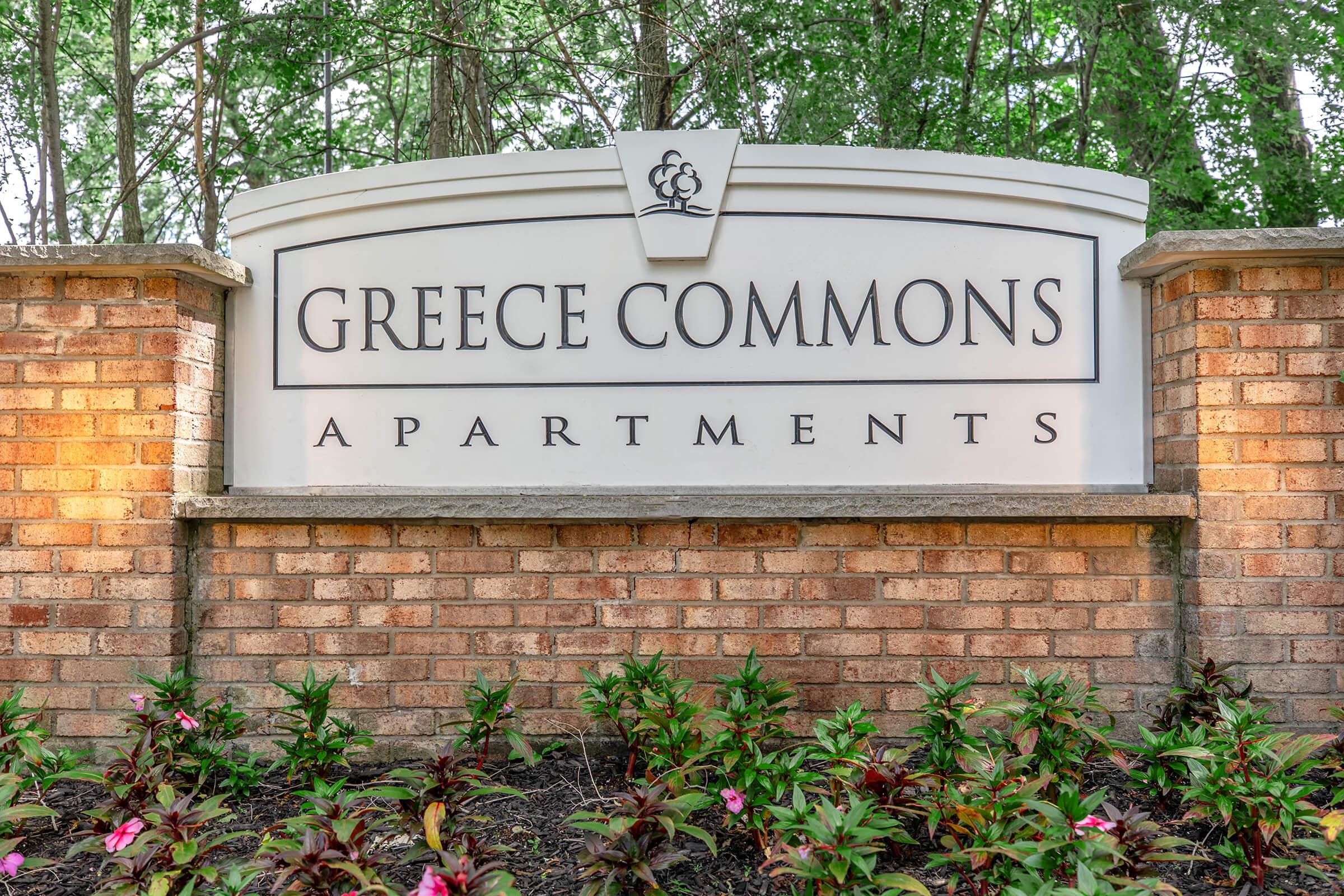 Sign for Greece Commons Apartments with a brick base and surrounded by greenery and flowers.