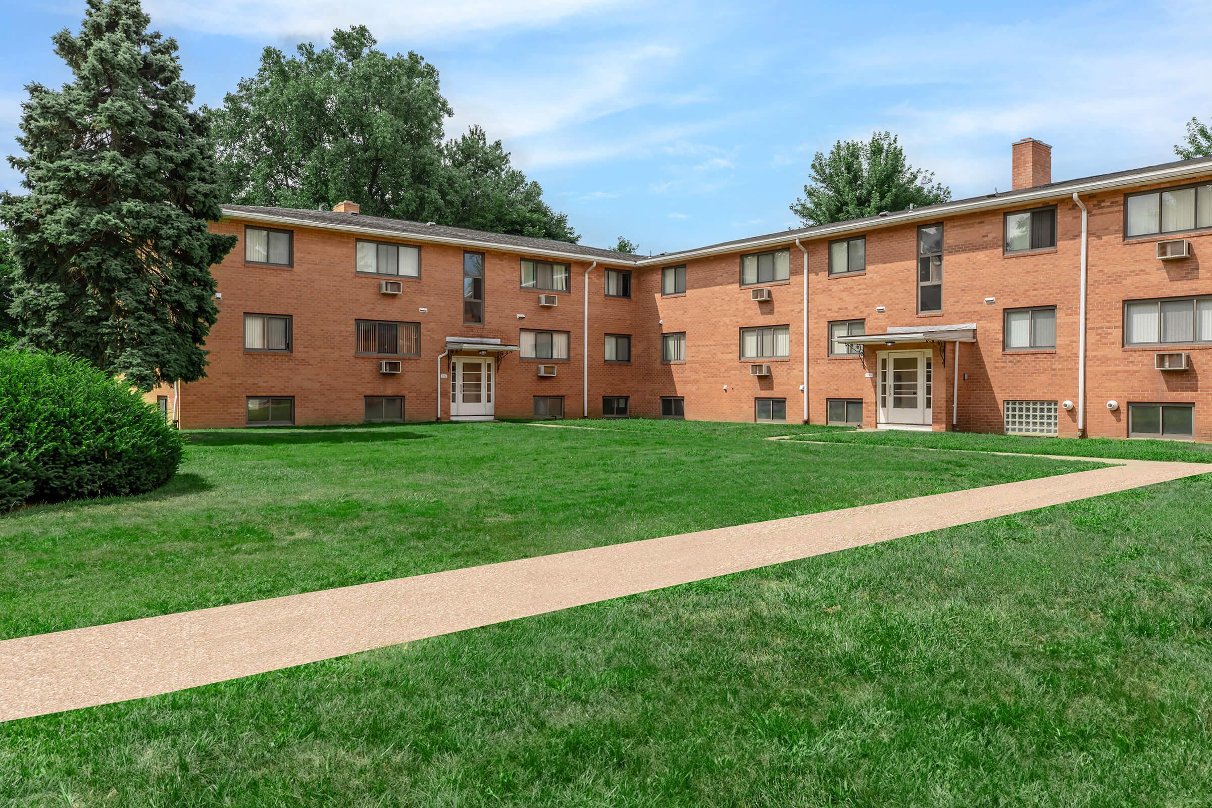 A view of a low-rise brick apartment building surrounded by a grassy area, with a pathway leading up to the entrance. Several windows and air conditioning units are visible, along with trees in the background.