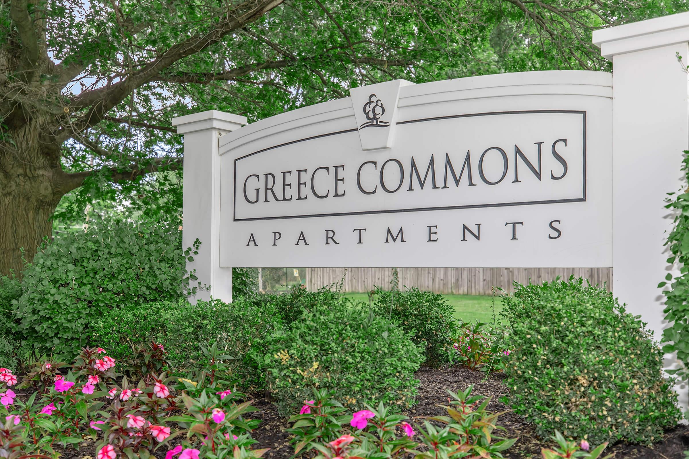 Sign for Greece Commons Apartments surrounded by lush greenery and flowering plants.