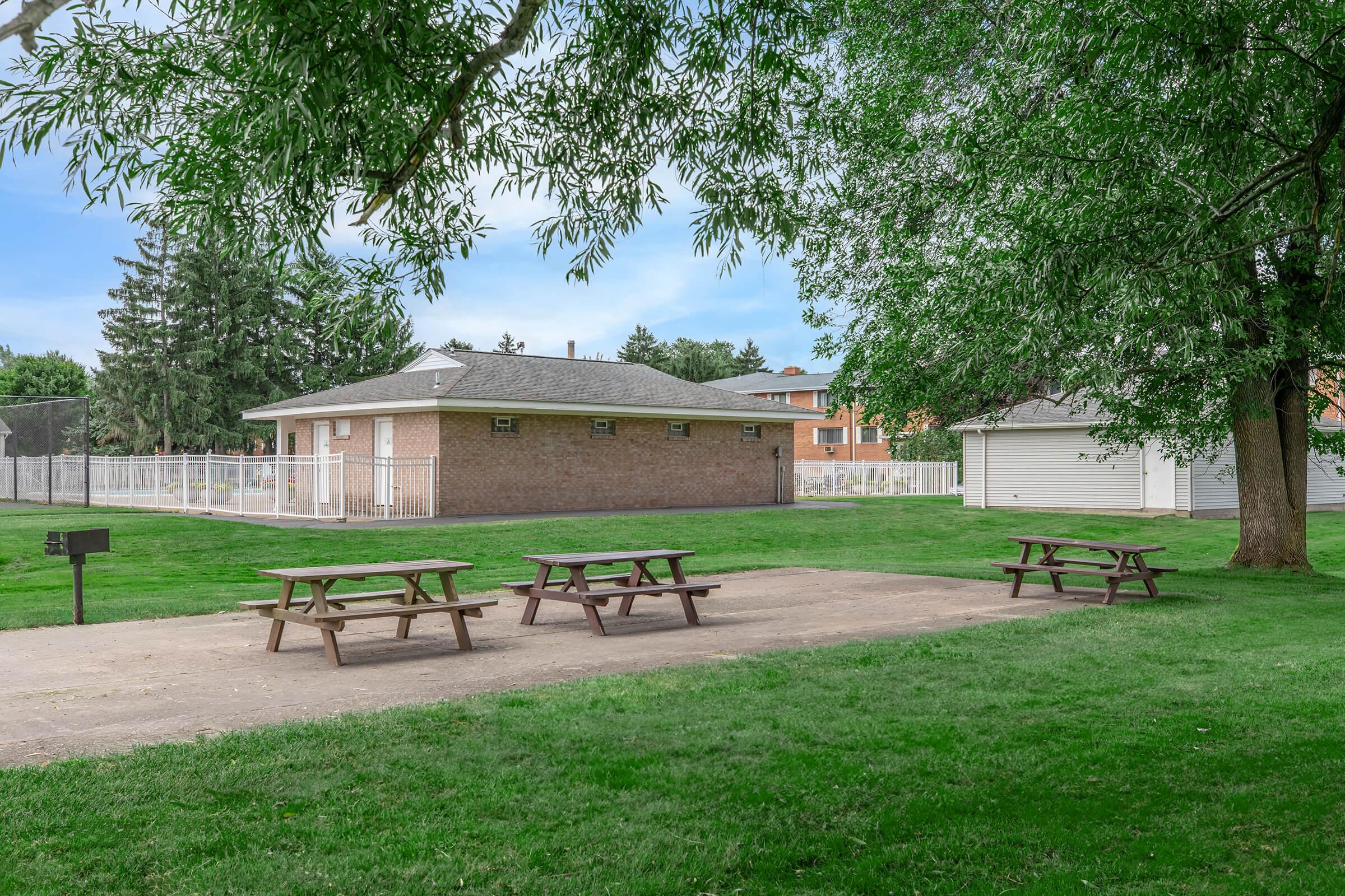 A grassy park area featuring several picnic tables, a brick building in the background, and trees providing shade. The scene includes a fence in the distance, suggesting a recreational space.