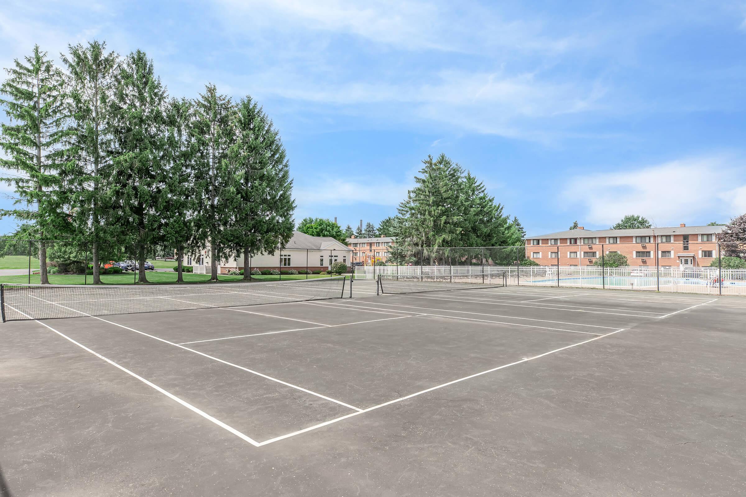 A tennis court with a gray surface and white lines, surrounded by tall green trees and residential buildings in the background. Clear blue sky overhead.