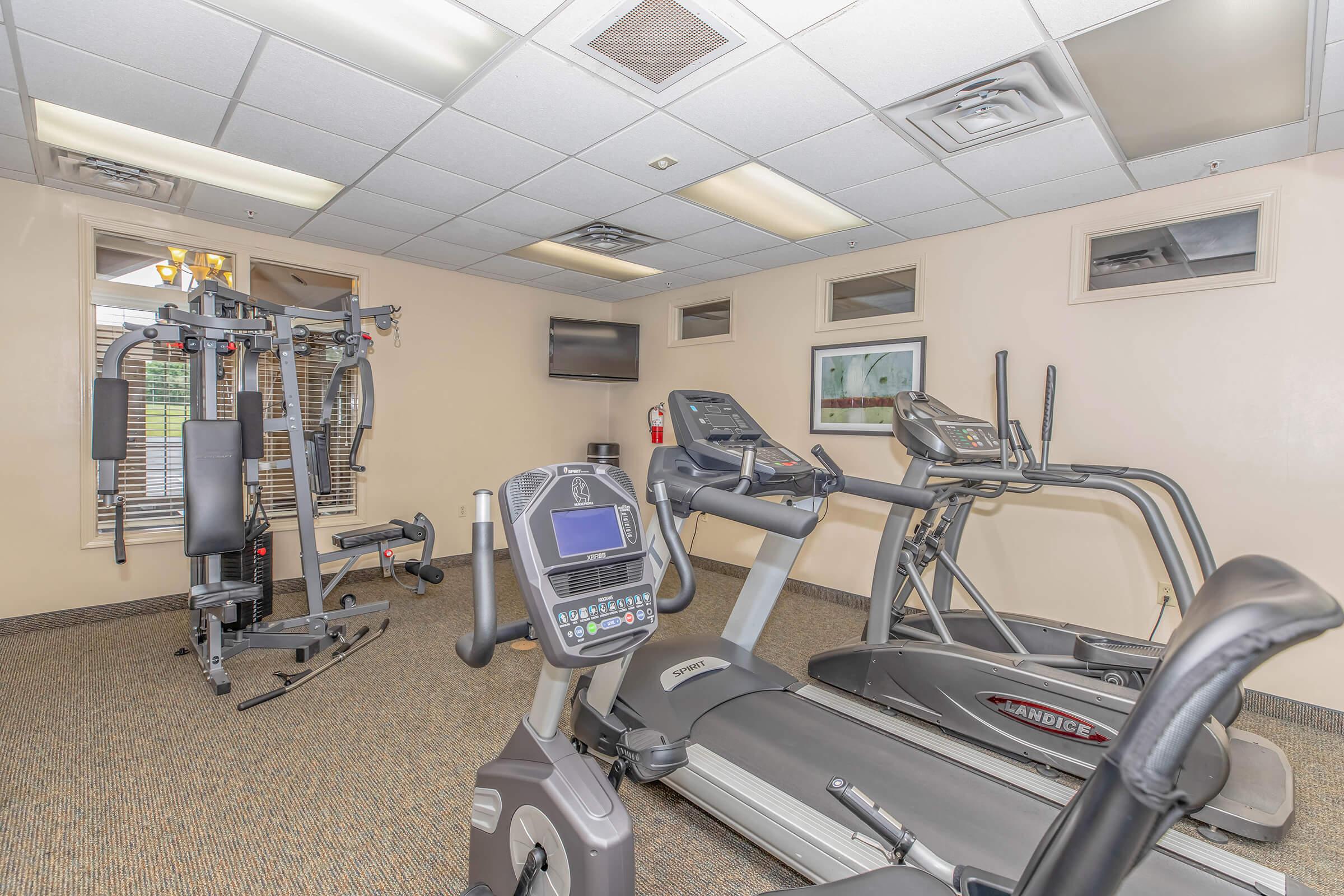 A modern gym interior featuring exercise equipment, including a treadmill, elliptical machine, and a weight machine. The room has soft lighting and is designed for fitness activities, with wall-mounted TVs and windows for natural light.