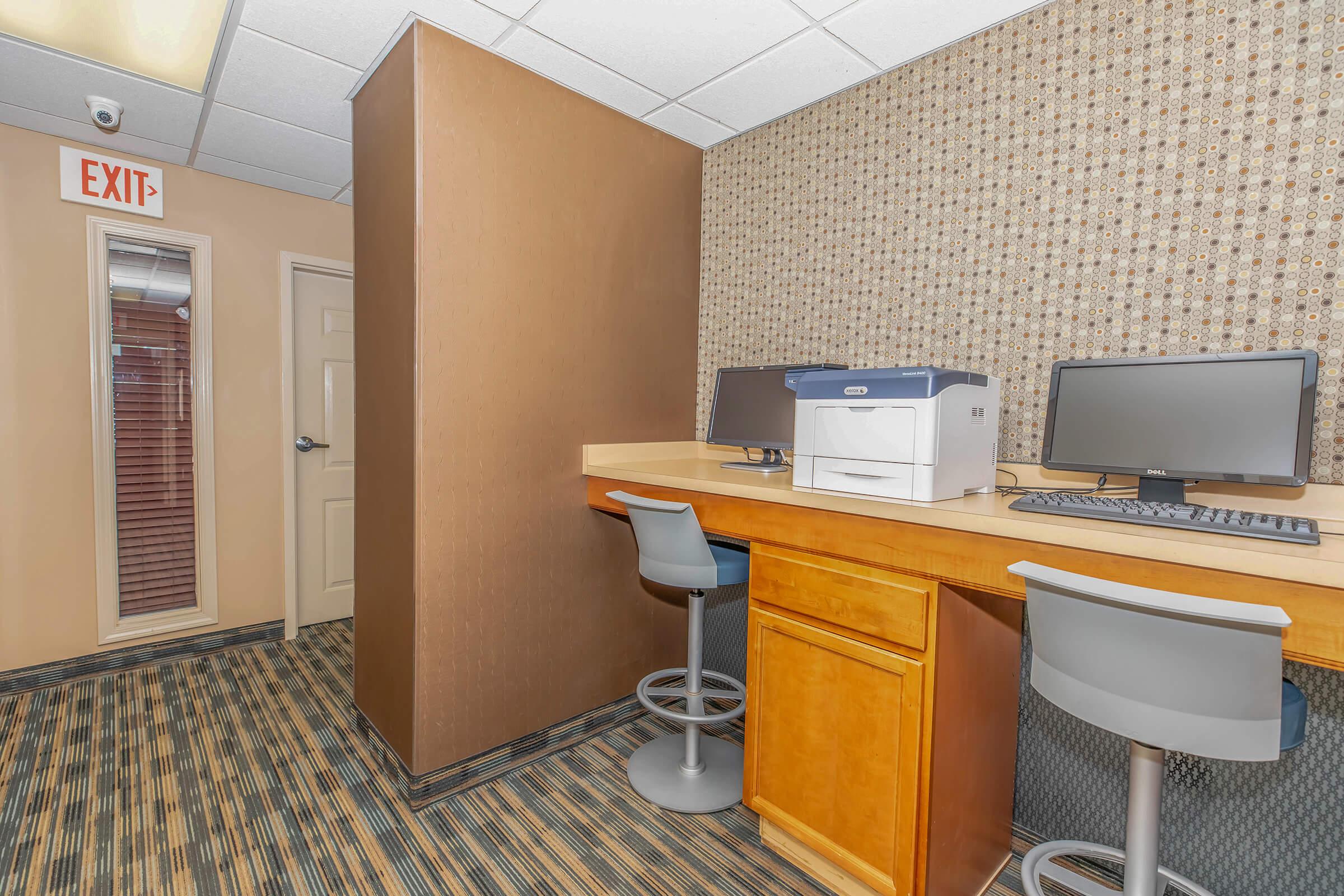 A small office space featuring a desk with two computers and a printer, along with two gray office chairs. There is an exit sign on the wall and a partition separating the workspace from an adjacent area. The carpet has a striped pattern, and the walls have a textured design.