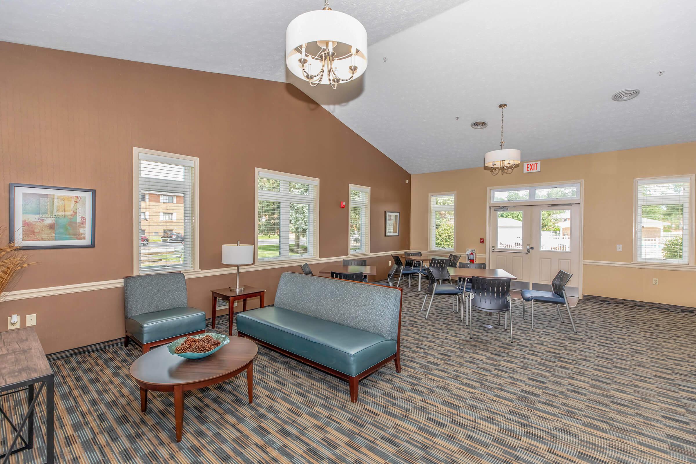 Interior view of a spacious common area featuring seating with modern furniture, a coffee table, and large windows. The walls are a warm brown tone, and the ceiling has decorative lighting. Textured carpet covers the floor, and there are dining tables in the background.