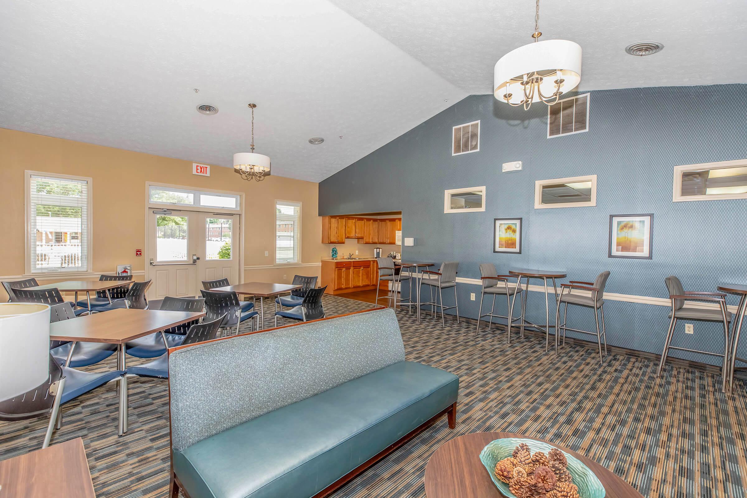 A cozy lounge area with tables and chairs, a small kitchen counter in the background, and light-colored walls featuring framed artwork. The space is well-lit with large windows and decorative lighting.