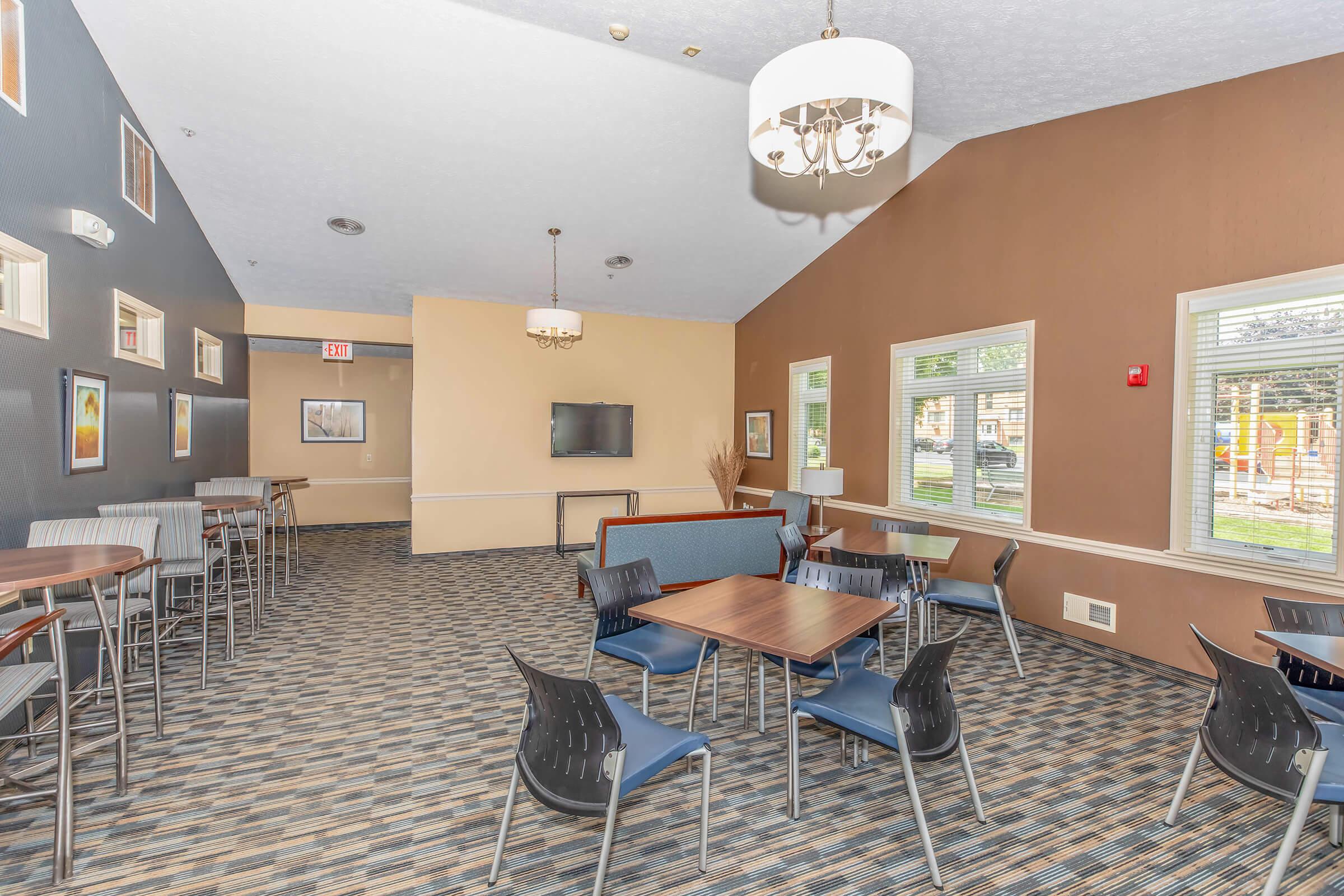 A spacious lounge area featuring a mix of tables and chairs, wall-mounted television, and large windows allowing natural light. The room has a modern design with neutral-colored walls and patterned carpeting.