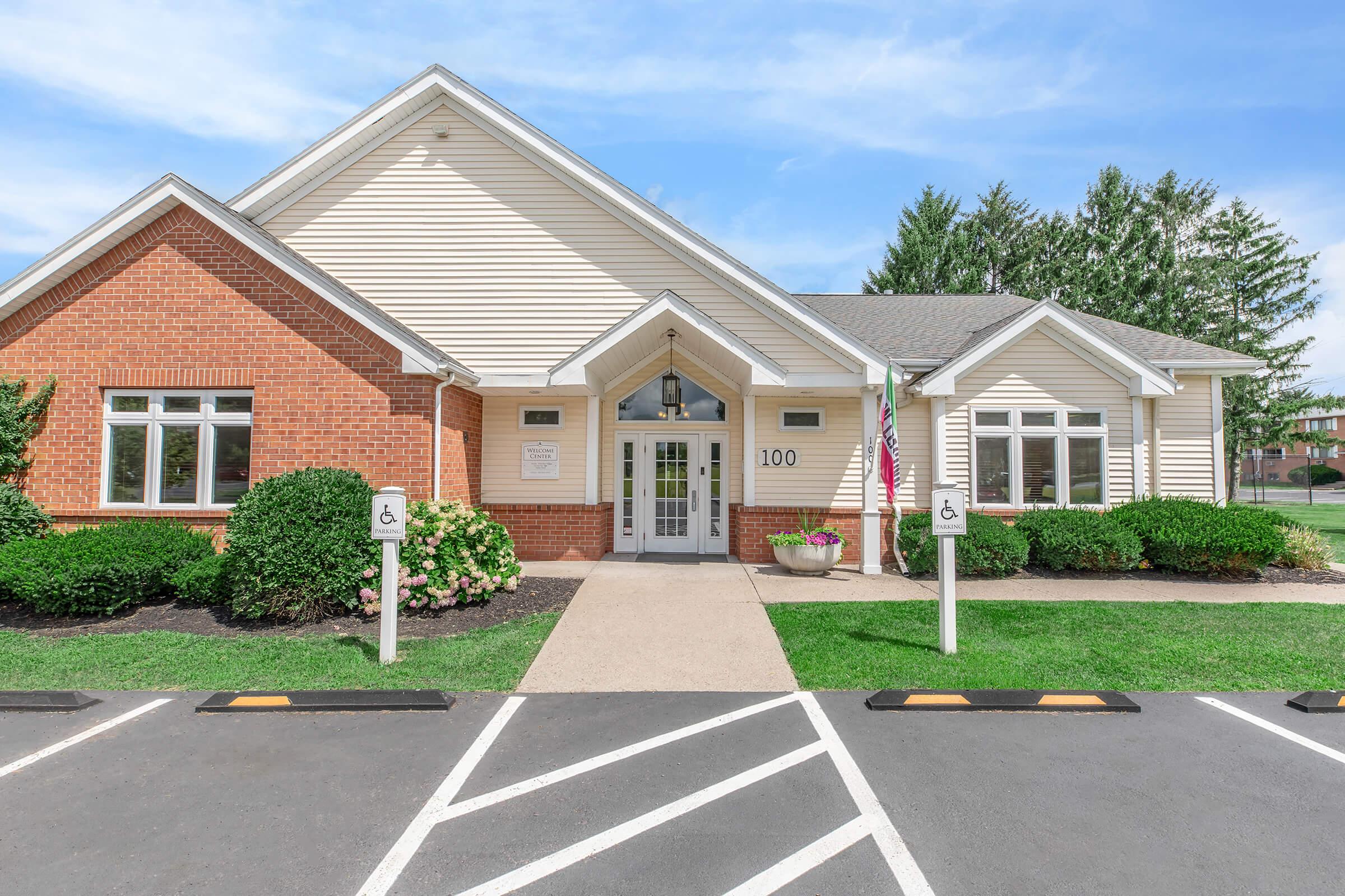 Single-story building with a light-colored exterior and red brick accents, featuring a central entrance with large glass doors, surrounded by well-maintained green landscaping and a parking area marked with accessible signs. The building has a welcoming appearance with a clear blue sky overhead.