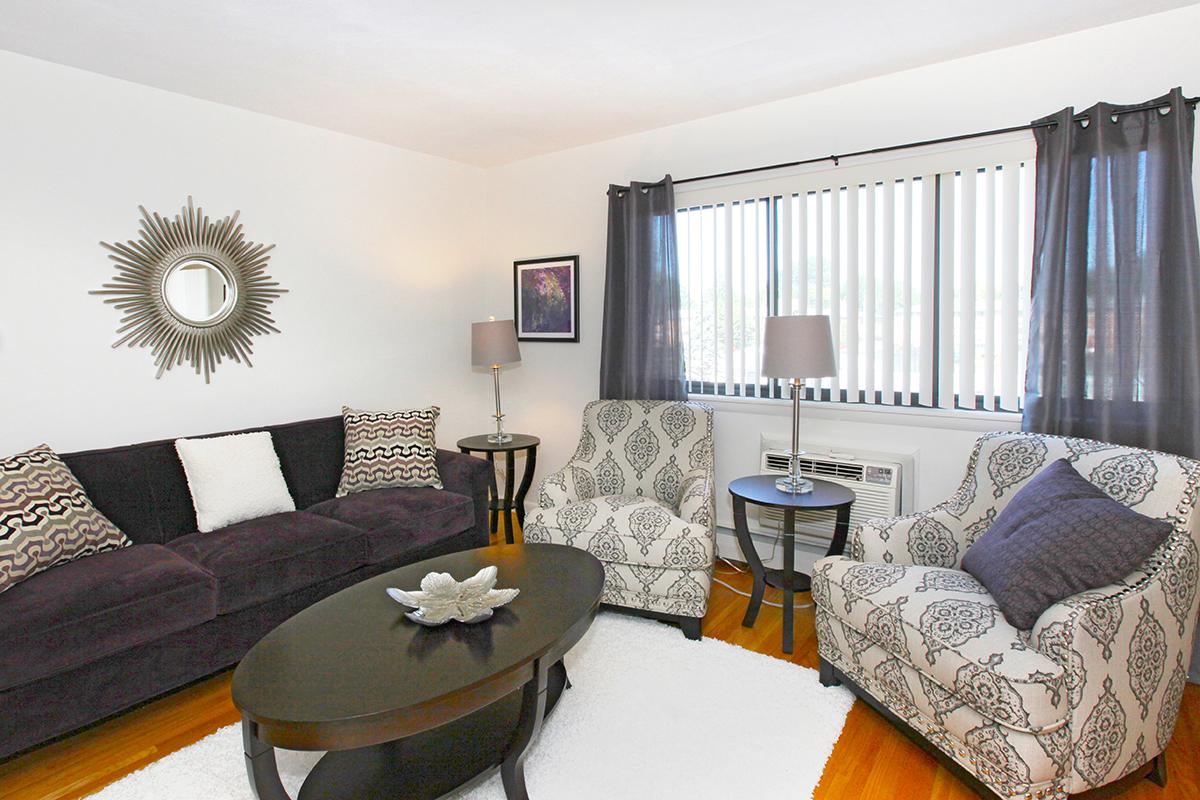 A cozy living room featuring a dark sofa with decorative cushions, two patterned armchairs, a round coffee table, and a mirror on the wall. The space has light-colored walls, dark curtains, and a small air conditioning unit beneath a window, creating an inviting atmosphere.