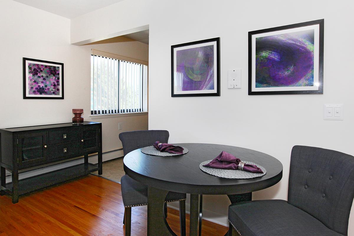 A modern dining area featuring a round black table set with purple napkins. Two gray chairs are positioned around the table. A dark sideboard is against the wall, and two framed abstract art pieces in purple tones are displayed. The room has natural light from a window with vertical blinds.