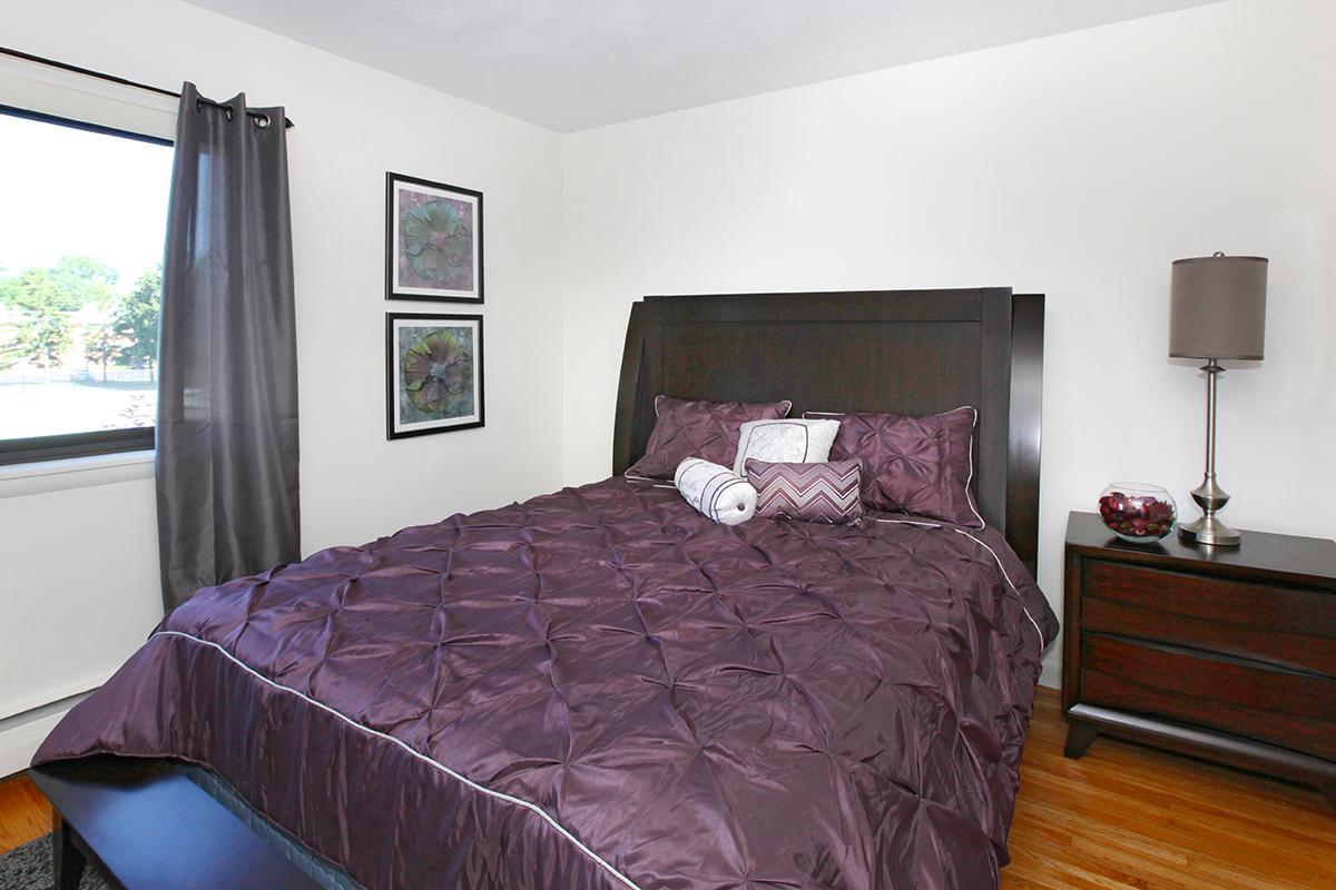 A cozy bedroom featuring a queen-sized bed with a dark purple quilt, accented by decorative pillows. There's a nightstand with a lamp beside the bed, and framed art on the wall. Natural light streams in through a window with gray curtains, creating a warm and inviting atmosphere.