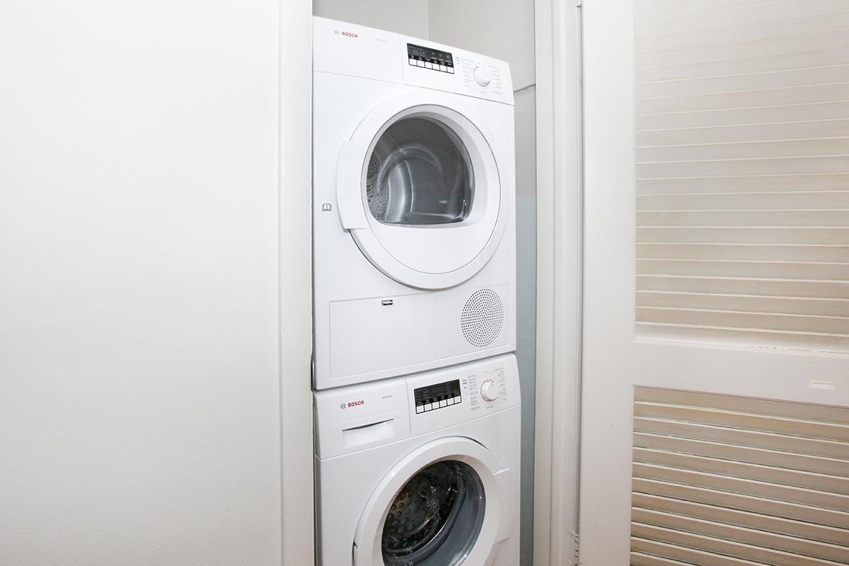 A stacked washer and dryer in a laundry room setting. The appliances are white and modern, featuring control panels with knobs and buttons. The background includes a partially visible door and wall, indicating a compact space for laundry appliances.