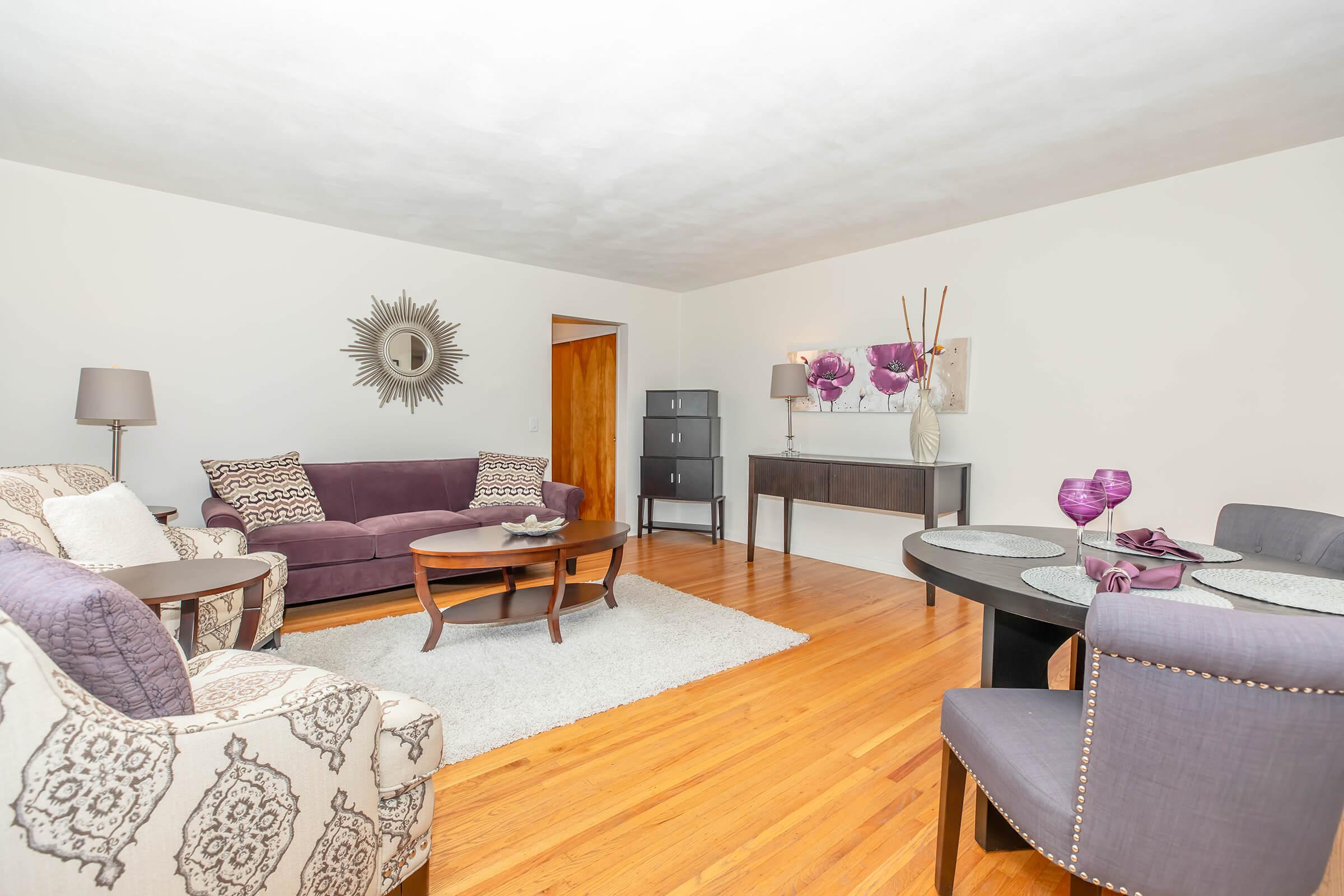 A cozy living room featuring a purple couch and patterned armchair, a round wooden coffee table, and a dining table with blue upholstered chairs. The space includes a decorative mirror, a sideboard, and floral accents, all set against light walls and hardwood flooring. A soft area rug adds warmth to the room.