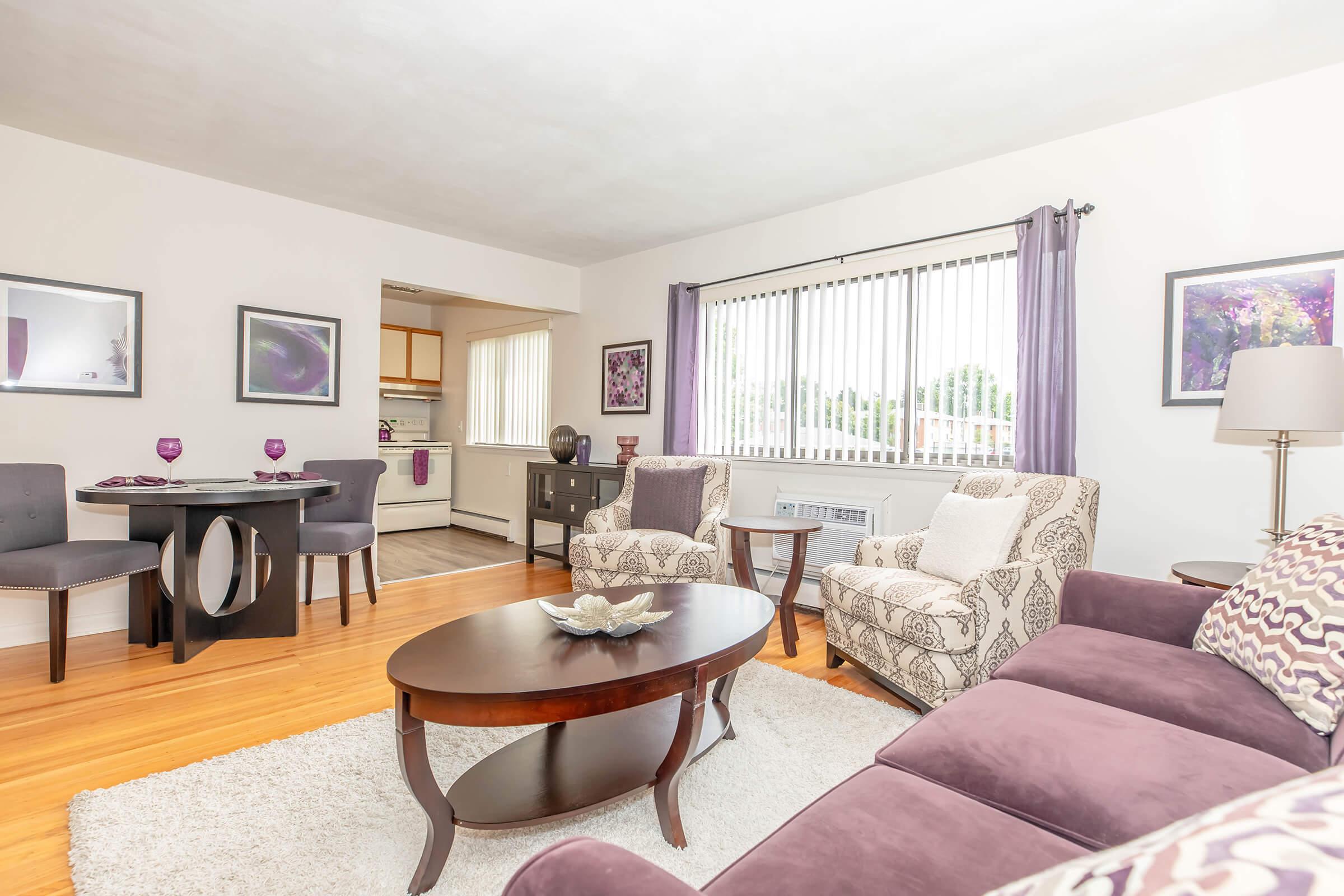 A cozy living room featuring a plush purple sofa, two patterned armchairs, and a round coffee table with a decorative bowl. There's a dining area with a small table set for two. Large windows provide natural light, and the walls are adorned with framed artwork. The wooden floor adds warmth to the space.