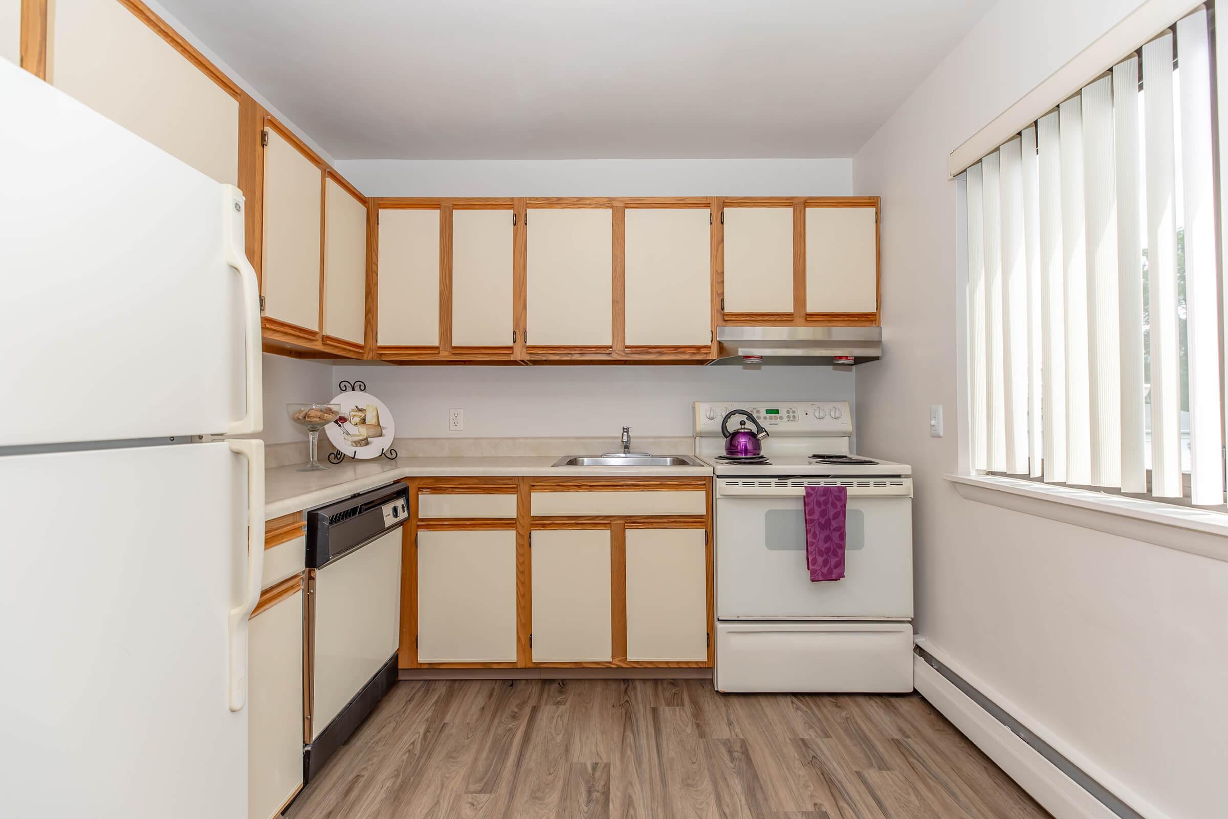 A modern kitchen featuring light-colored cabinets with wood trim, a white refrigerator, a sink, a stove with an oven, and a dishwasher. The countertop is beige, and the flooring is a light wood grain. The room has vertical blinds on the window, allowing natural light to enter.