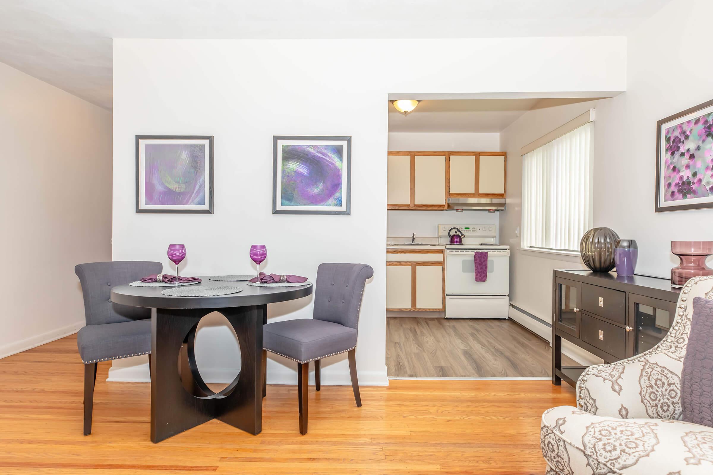 A modern dining area features a round black table with two gray chairs, set with purple glasses and plates. In the background, a kitchen with light-colored cabinets and an oven is visible, along with two abstract art pieces in purple tones hanging on the wall. The flooring is hardwood.