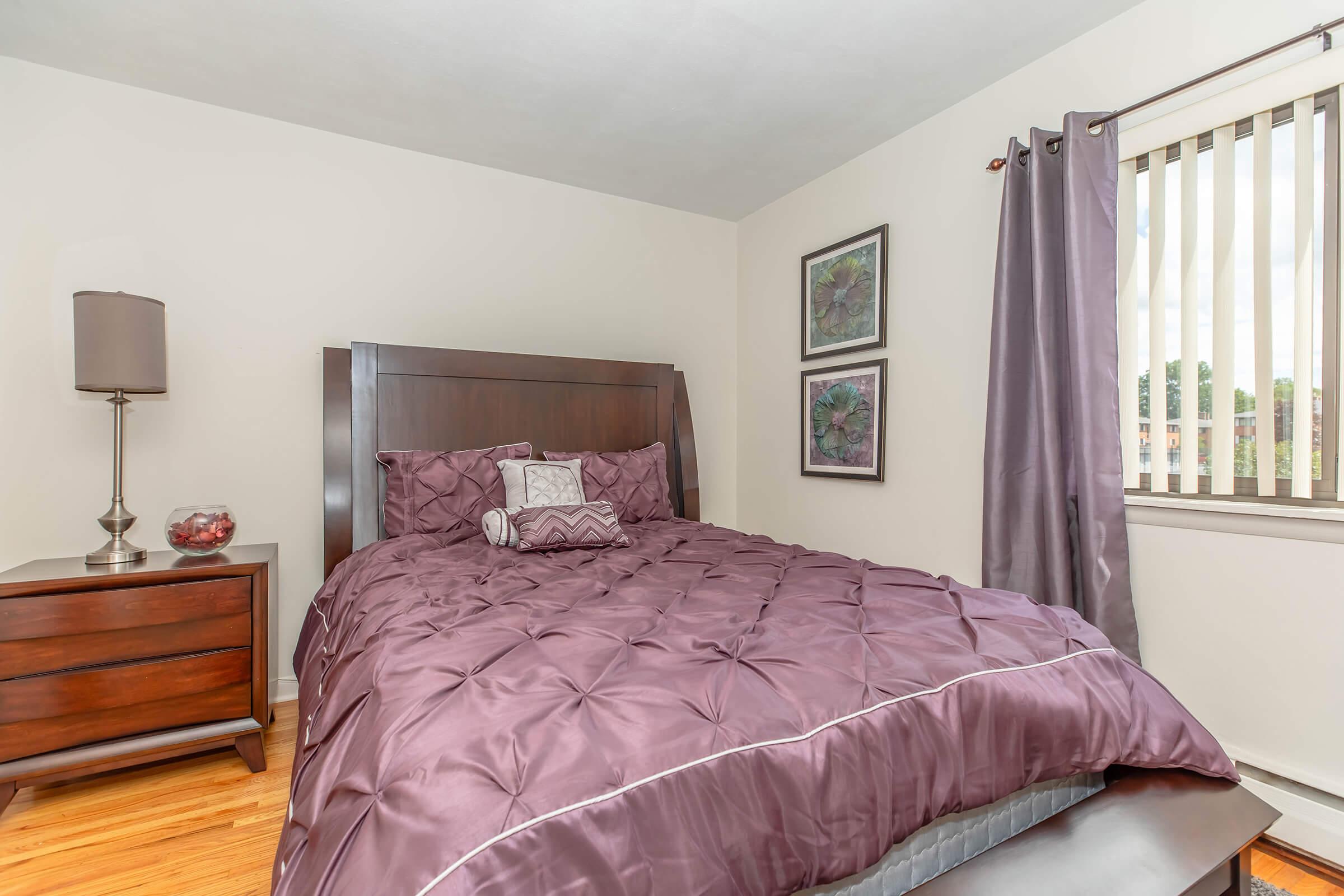 A cozy bedroom featuring a dark wooden bed with a plush purple comforter, a decorative pillow, and matching bedside table with a lamp. The room includes two framed artworks on the wall and light-colored curtains with vertical blinds, providing a warm and inviting atmosphere.