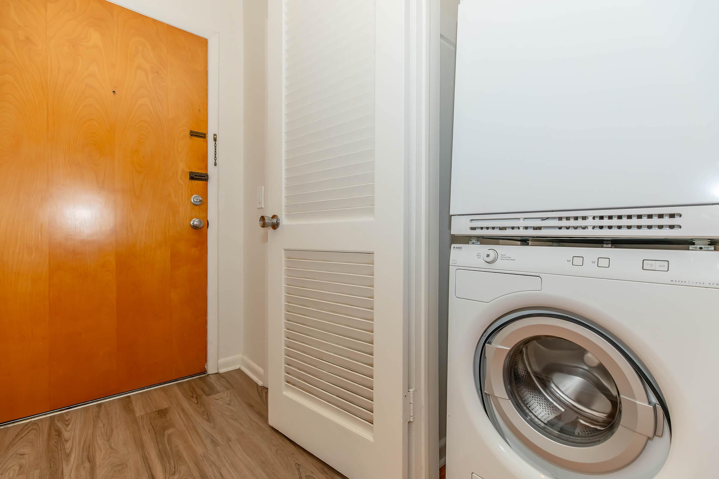 A view of a small entryway featuring a wooden front door with a peephole, beside a white stacked washing machine and dryer. The wall next to the door includes a white louvered door for closet space, and light brown flooring creates a warm atmosphere in the space.