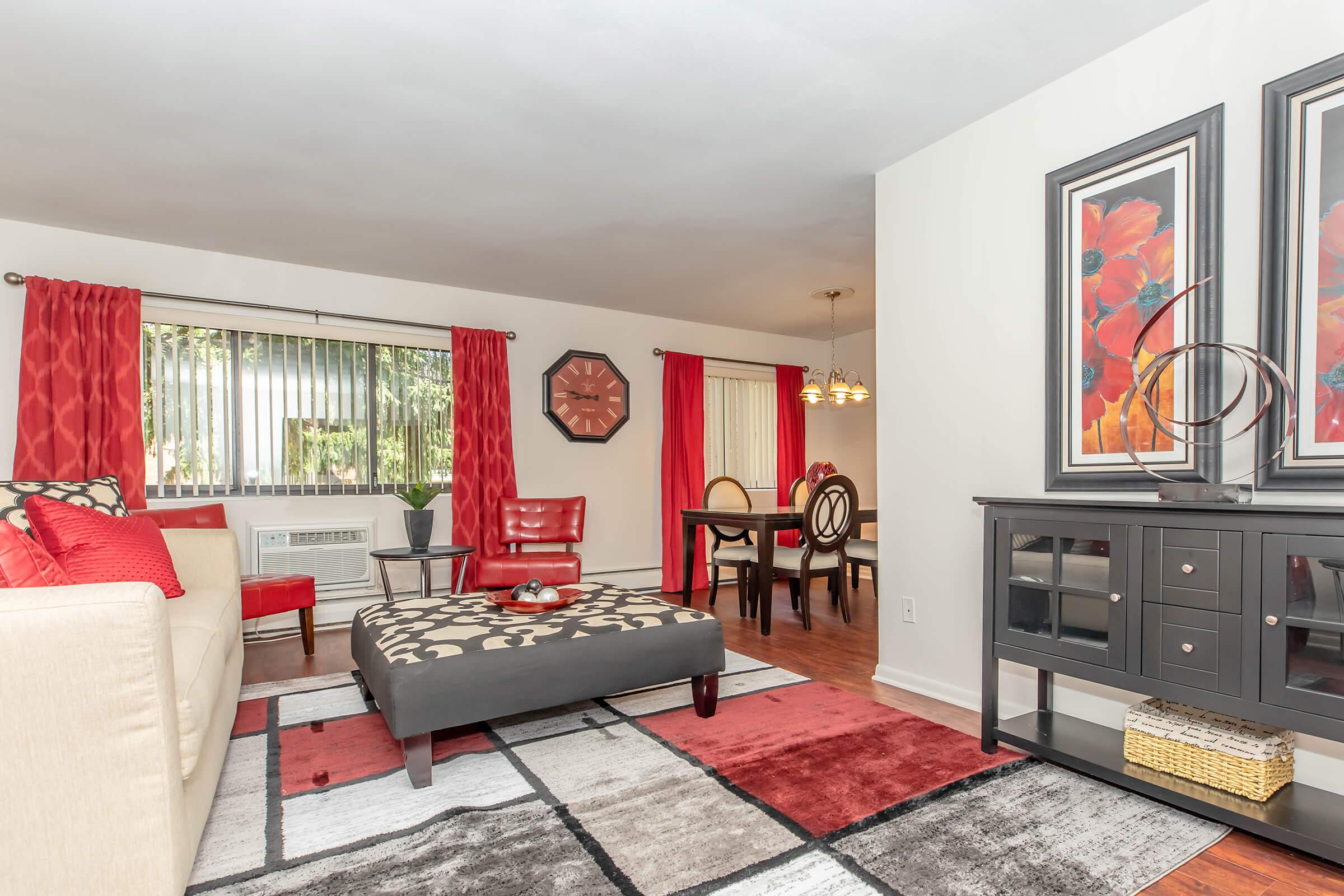 A stylish living room featuring a cream-colored sofa with red and patterned accents, a modern coffee table, and a decorative rug. Large windows with red curtains allow natural light. A dining area is visible in the background, showcasing a round table and chairs, alongside elegant wall art.