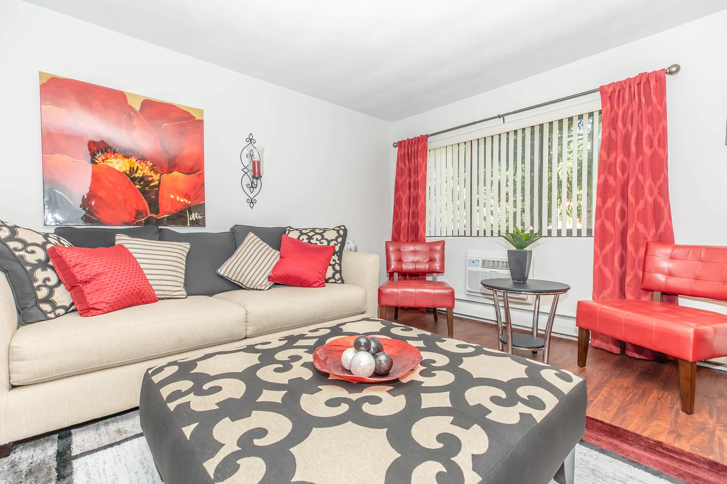 A modern living room featuring a beige and gray sofa adorned with red and black decorative pillows. A large, vibrant red floral artwork hangs on the wall. There is a circular coffee table with decorative balls on it, flanked by two red chairs. Light streams through partially open blinds, enhancing the cozy atmosphere.