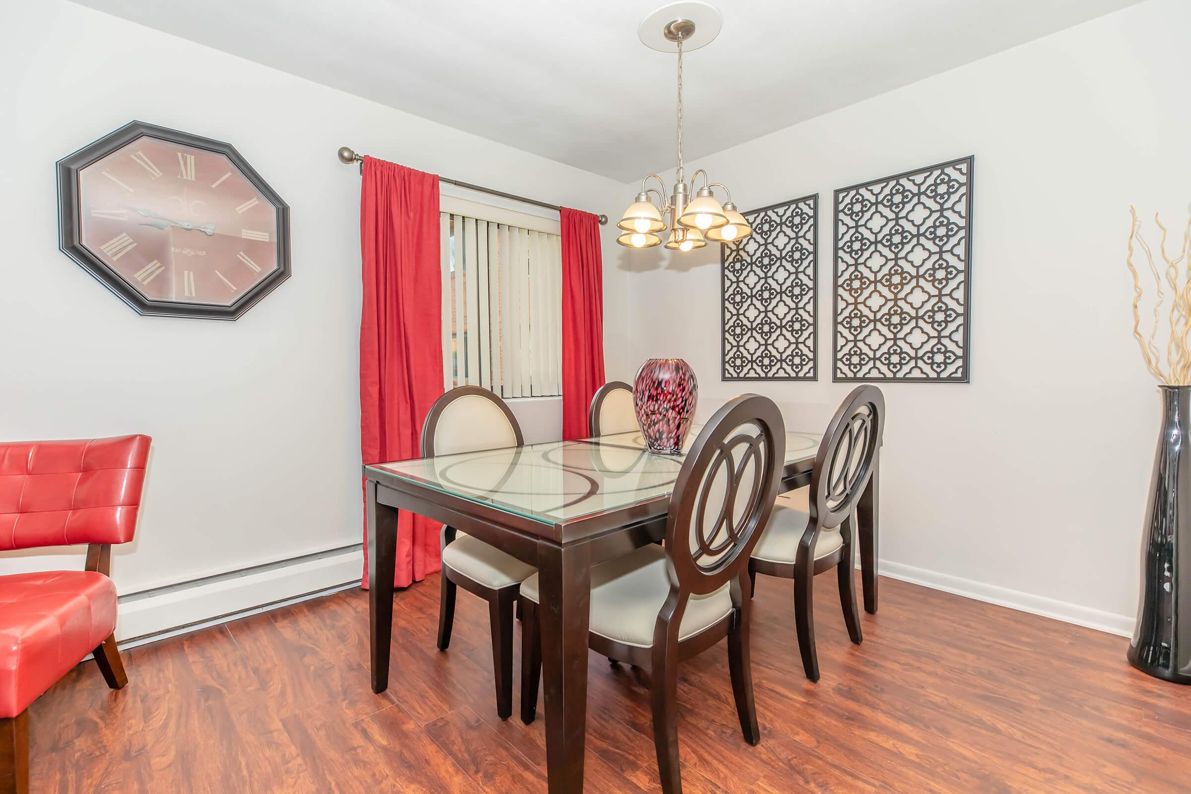 A modern dining room featuring a glass-top table with six upholstered chairs. There is a large wall clock and two decorative framed pieces on the wall. Red curtains frame a window, and a tall, slender vase stands in the corner. The floor is wooden, adding warmth to the space.