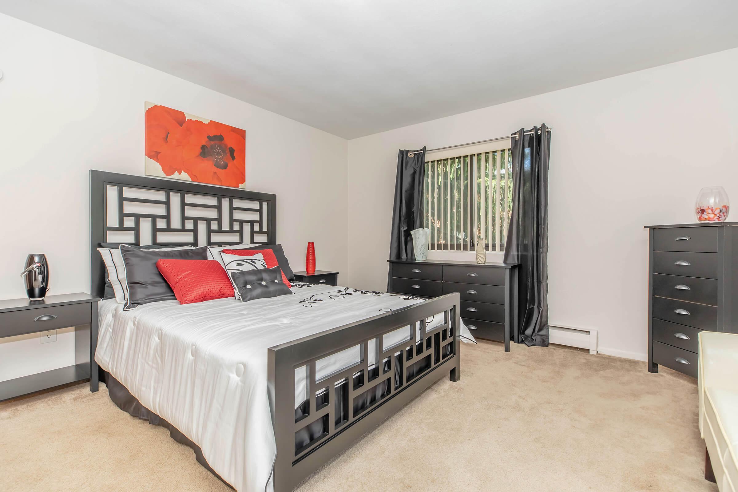 A well-decorated bedroom featuring a stylish black bed frame with a geometric headboard. The bed is adorned with gray and red bedding. The room includes a matching dresser, two nightstands, and large windows with blinds, allowing natural light to flow in. Bright artwork on the wall adds a pop of color.