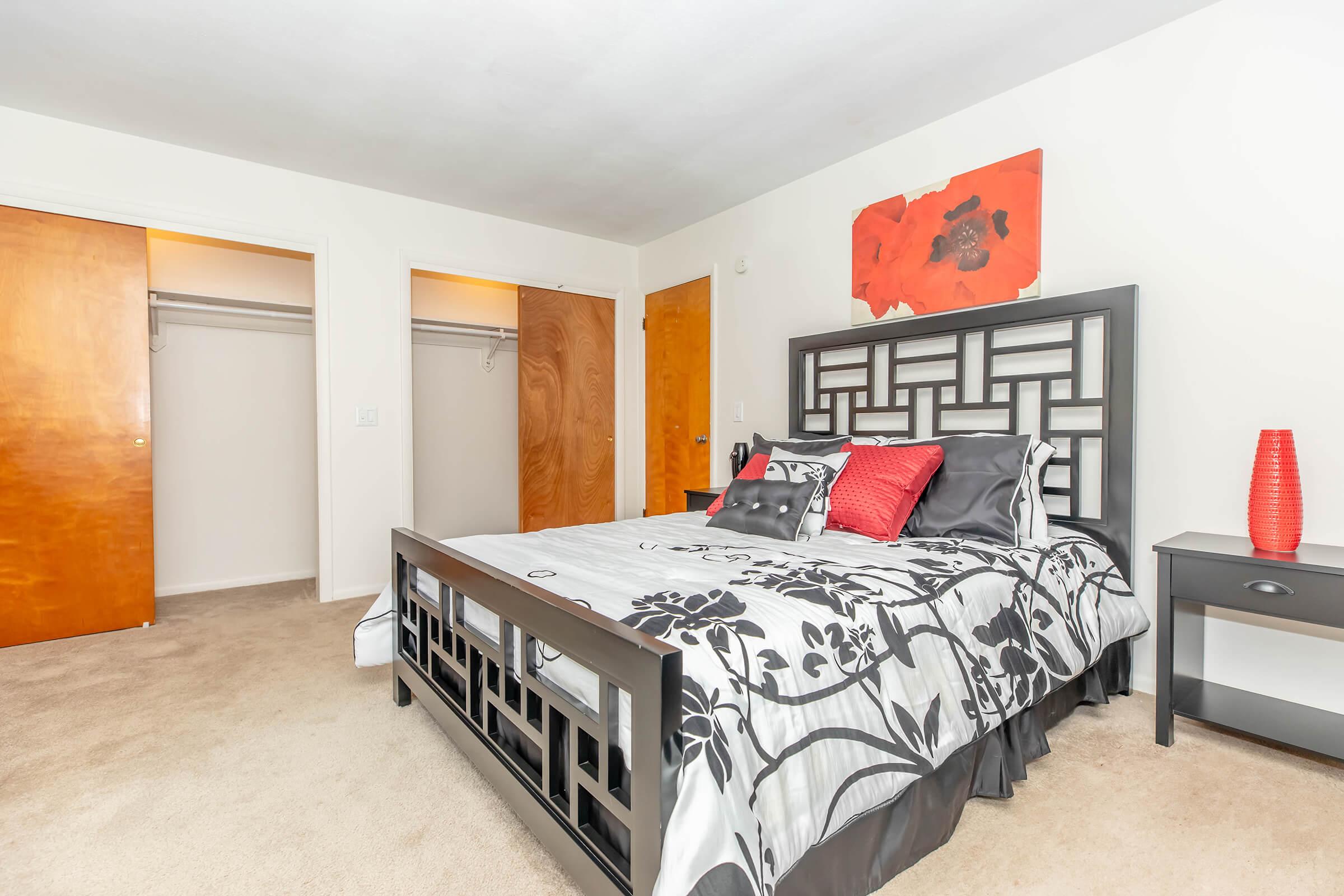 A cozy bedroom featuring a black and white floral bedding set on a queen-sized bed. The room has a minimalistic design with a red accent pillow and a decorative red vase on a bedside table. Two wooden closets are visible in the background, and the walls are adorned with a simple red artwork. Soft beige carpet covers the floor.