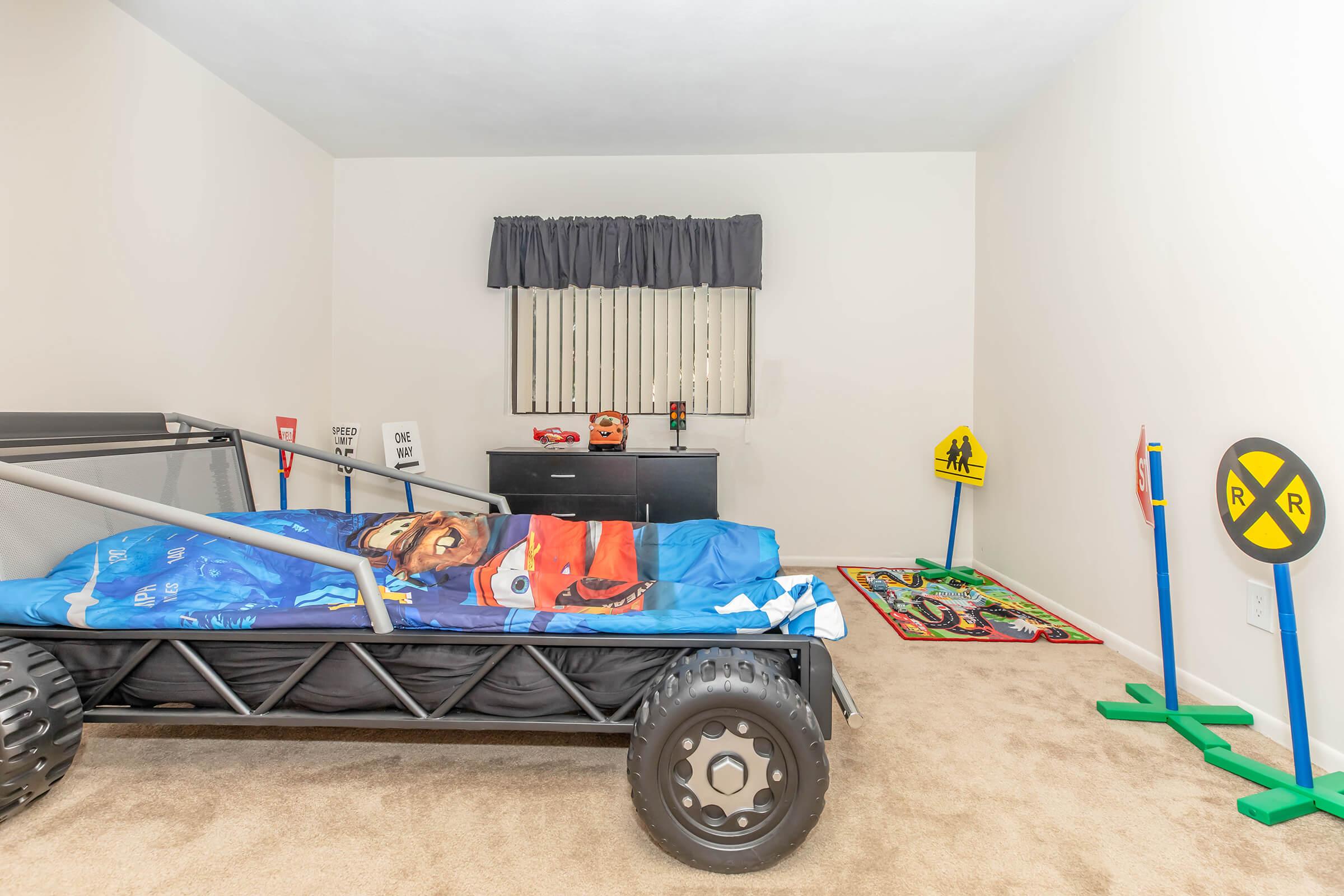 A child's bedroom featuring a race car-themed bed with blue bedding, a dark dresser, and various colorful toy traffic signs. The room is carpeted with a light brown shade and has a window covered by grey curtains. A colorful rug is visible on the floor, adding to the playful atmosphere.