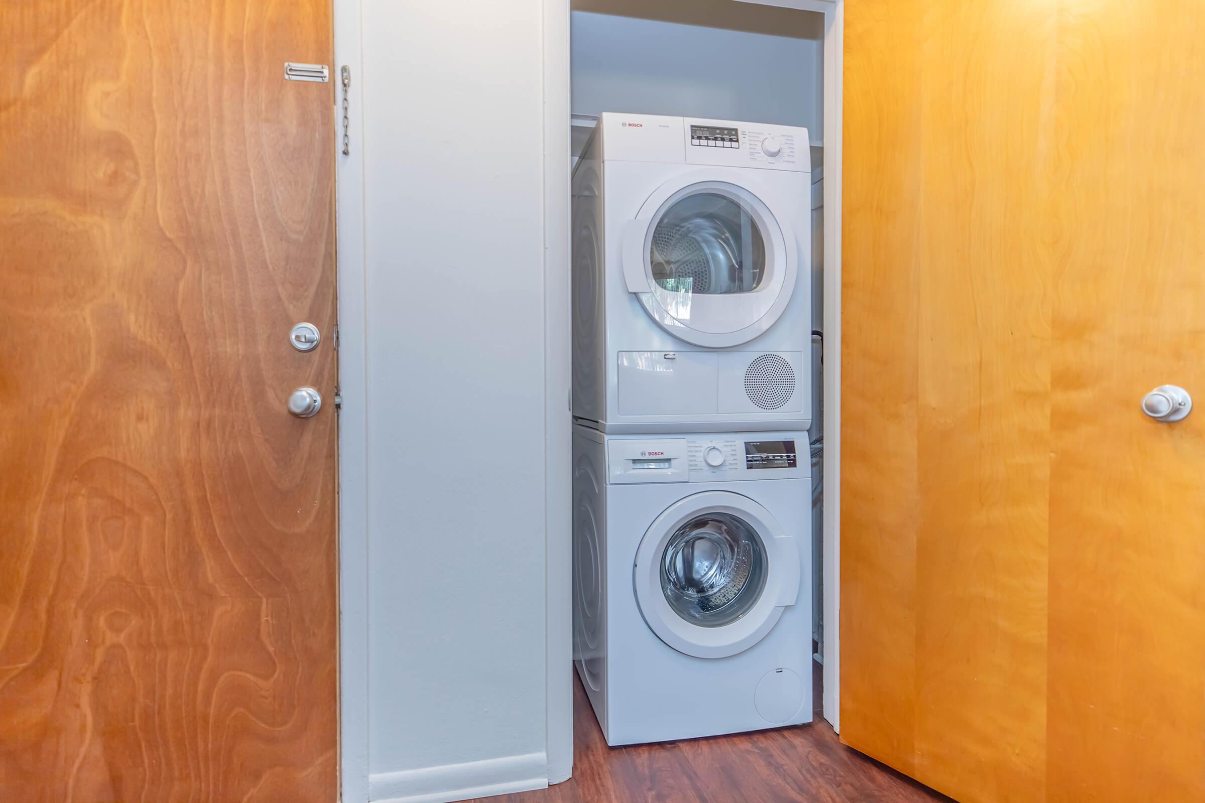 A compact laundry area featuring a stacked white washer and dryer unit located between two wooden doors. The space has light-colored walls and a wooden floor, creating a clean and modern look.