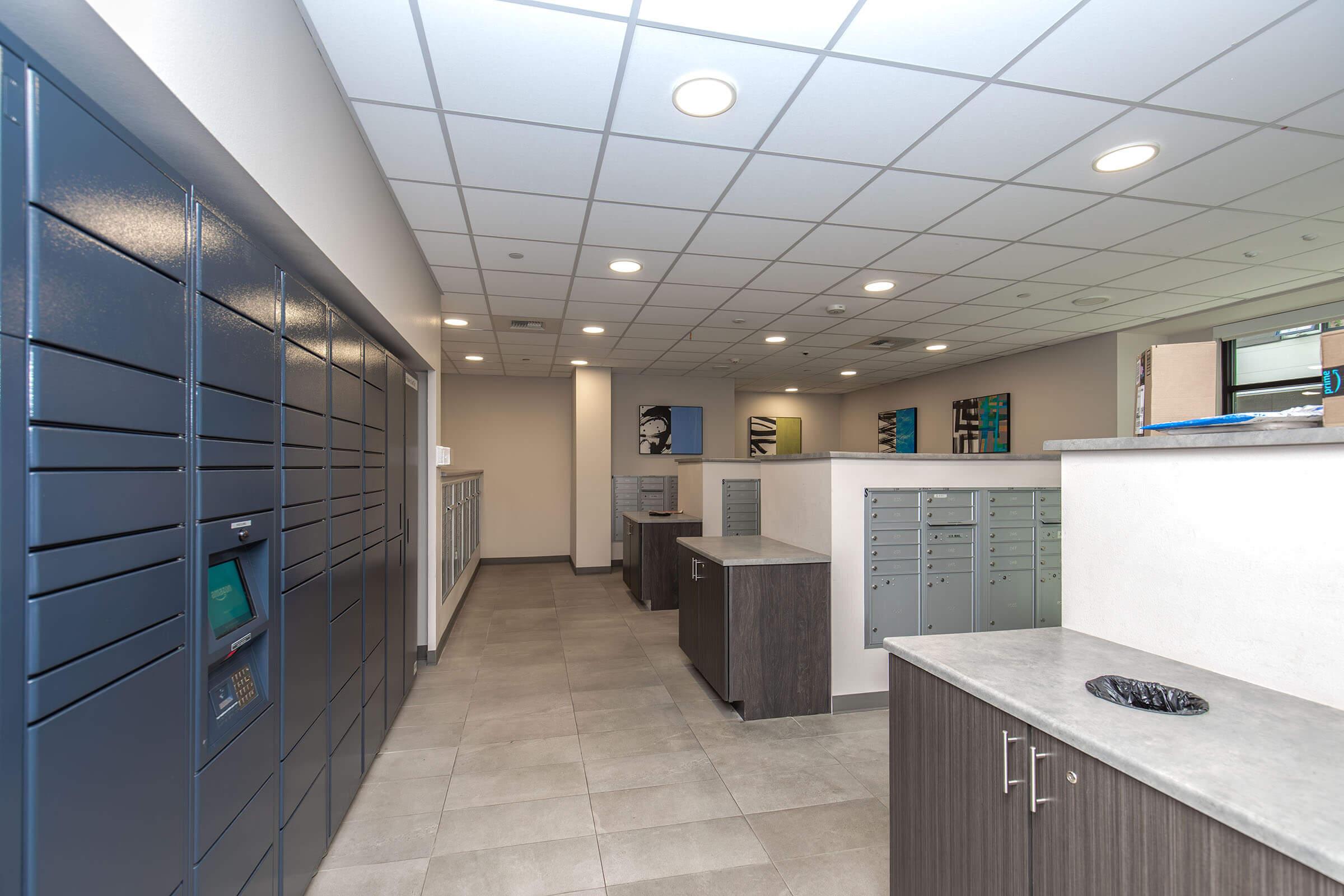 A spacious interior of a modern mailroom or package delivery area featuring multiple gray lockers, countertop workspaces, and wall-mounted artwork. The flooring is tiled, and the ceiling has recessed lighting, creating a bright and organized environment for managing mail and packages.