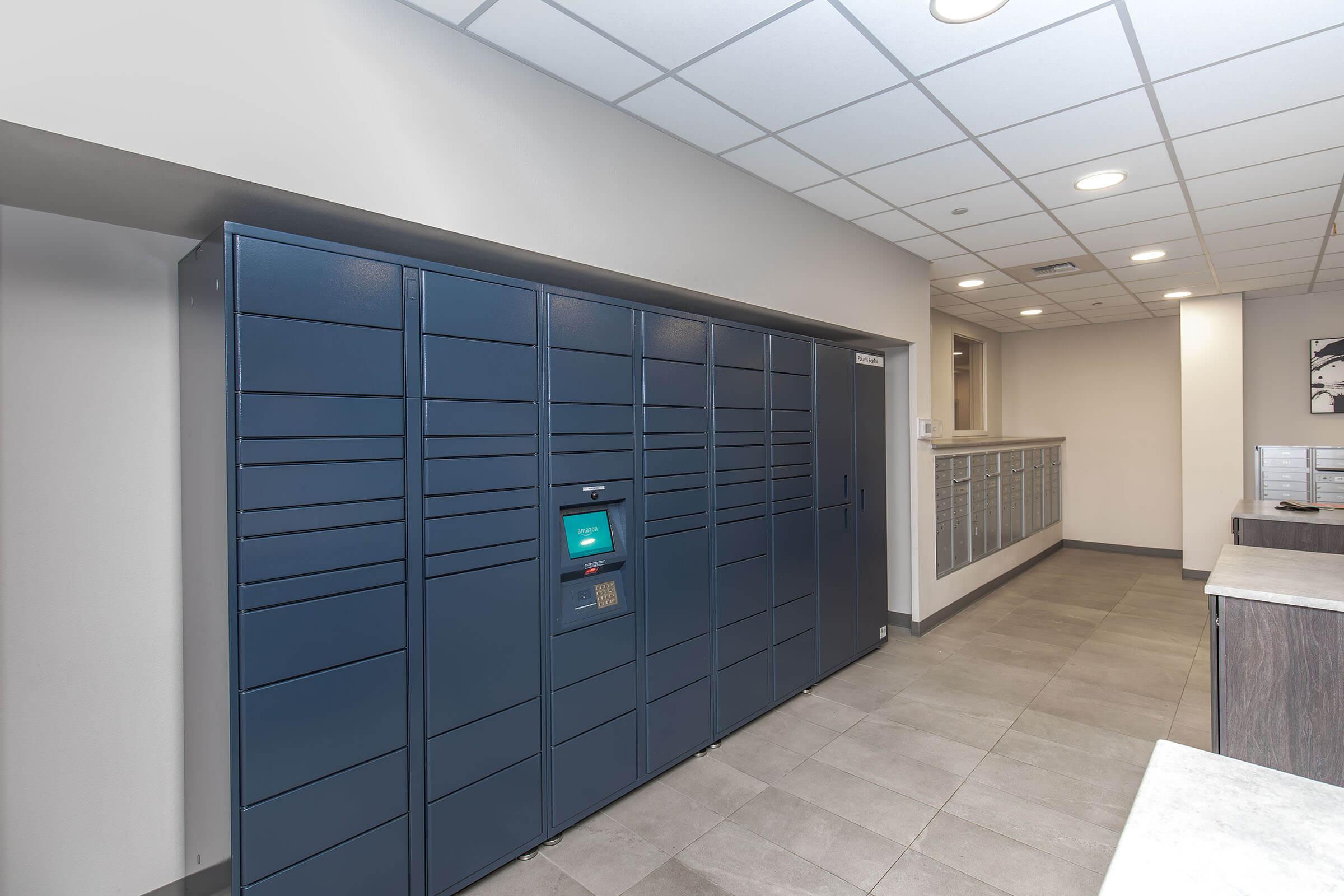 A modern, sleek package locker system is installed in a well-lit interior space with gray tile flooring. The locker unit is dark blue and features a digital touchscreen for user interaction. Additional mailboxes line the wall behind the lockers, and the overall decor appears contemporary and functional.