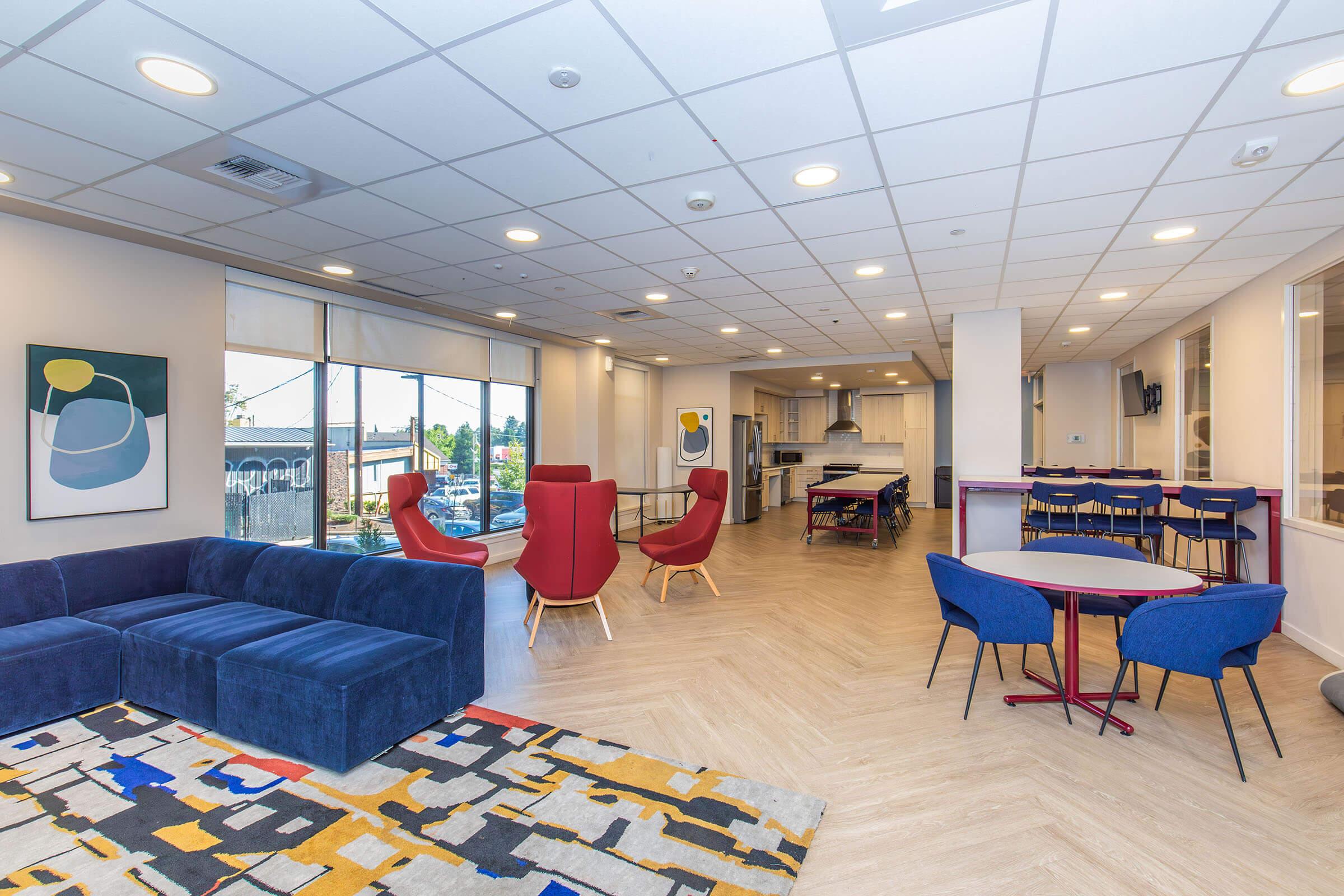 A modern, spacious common area featuring a blue sofa, red accent chairs, and a patterned rug. There are tables and chairs for dining or meetings, large windows allowing natural light, and abstract artwork on the walls, creating a welcoming and vibrant atmosphere.