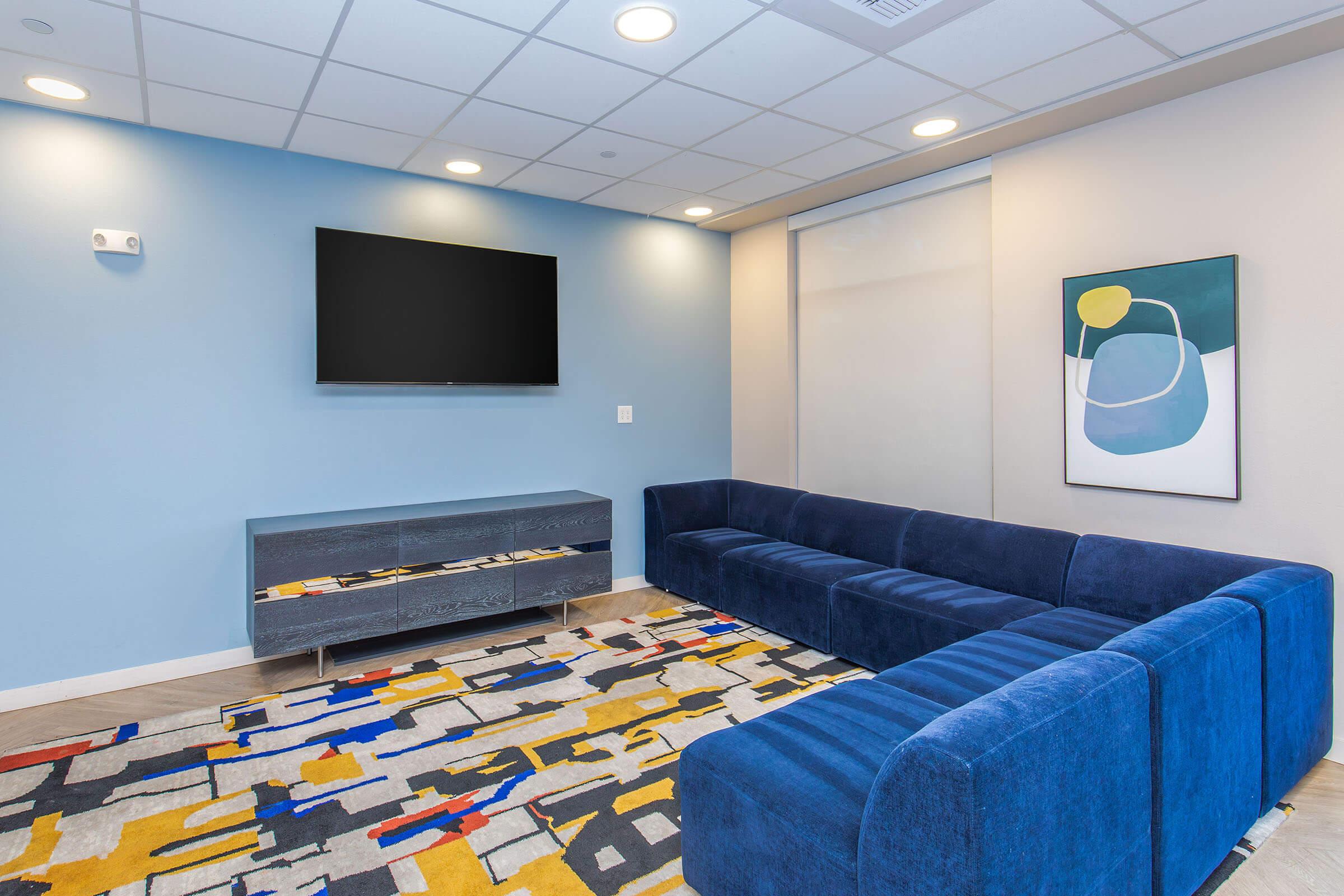 A modern living room featuring a blue sectional sofa, a flat-screen TV mounted on a light blue wall, and a stylish rug with abstract patterns in yellow, red, and blue. A minimalistic wall art piece adds a contemporary touch to the space.