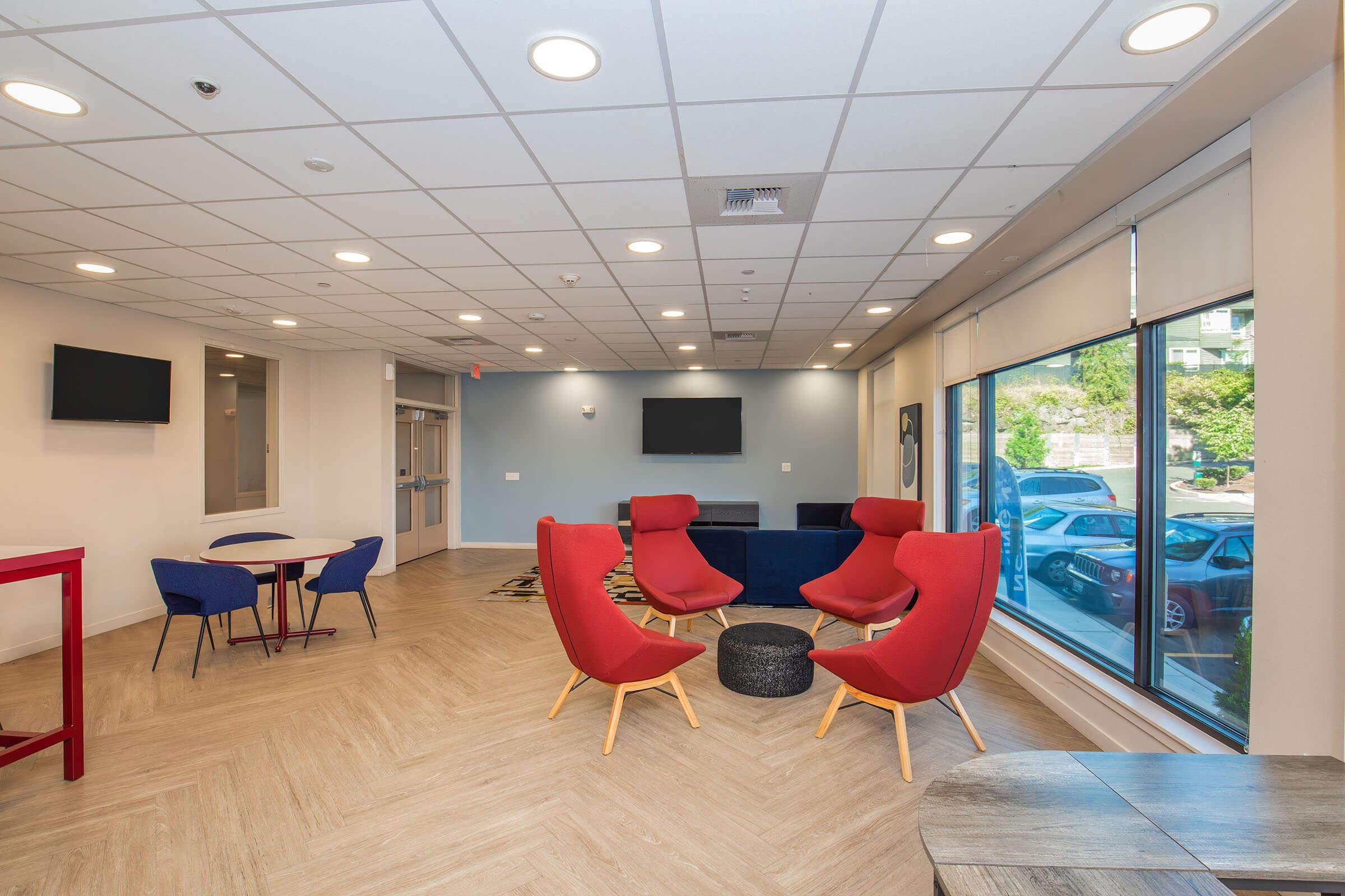 A modern lounge area featuring red armchairs, a round black coffee table, blue dining chairs, and a small round table. The space has large windows allowing natural light, a light-colored tiled floor, and flat-screen TVs mounted on the walls, creating a welcoming atmosphere for relaxation or socializing.